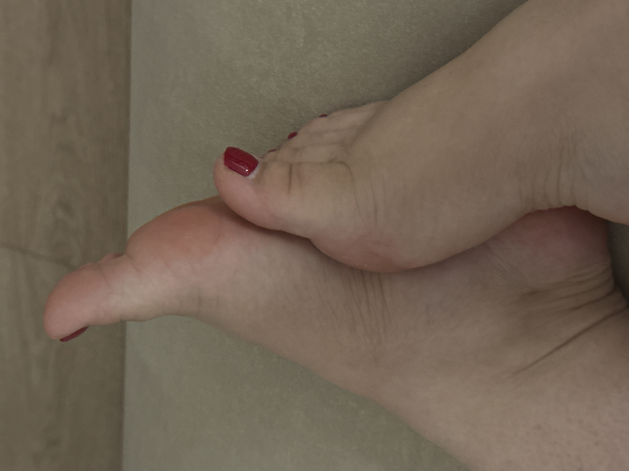 The image shows a close-up view of two human feet, with one foot resting on top of the other. Both feet have toenails painted in a dark red color. The skin appears fair and smooth with some visible texture and wrinkles, particularly around the ankles. The background is a neutral, textured surface, possibly a mat or carpet, with a hint of a wooden-like texture at the top of the frame. There are no discernible elements that place the image in Saratov, Russia, or any specific urban location.