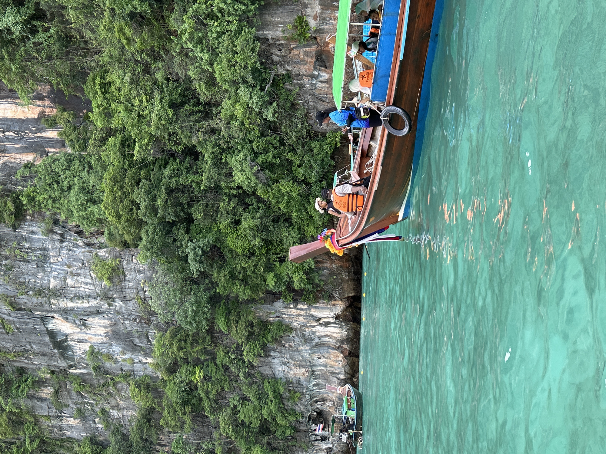 The image shows a longtail boat navigating through clear turquoise waters near a limestone cliff face covered in lush green foliage. On the boat, several individuals are visible. One person, wearing a blue shirt and dark pants, stands towards the rear, possibly operating the boat. Another person in an orange life jacket is seated towards the front. Several other passengers, including a child wearing a white hat and an orange life jacket, are also on board. A second, smaller longtail boat with a green canopy is also in the water to the right, carrying more passengers. In the background, a Thai flag is visible on the smaller boat. The cliffs are sheer and rocky, with vegetation growing out of crevices. The water is calm with gentle ripples, reflecting the light. The scene suggests a typical tourist activity in the waters around Ao Nang, Thailand, known for its dramatic karst landscapes and beautiful beaches.