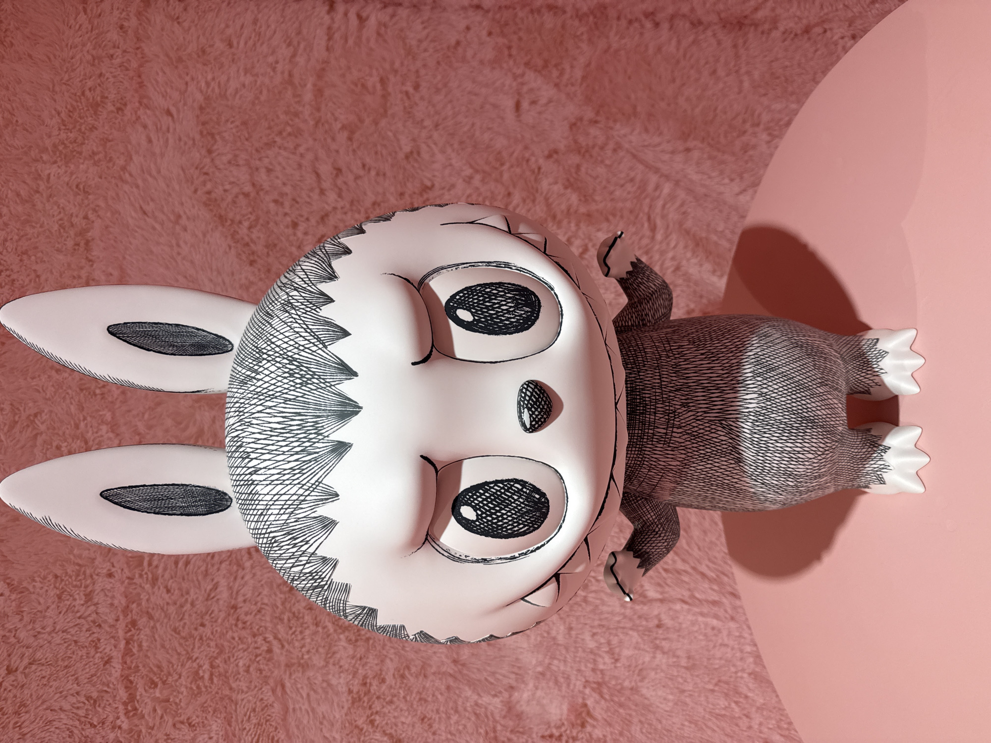 The image shows a close-up of a stylized, anthropomorphic toy figure with large, expressive eyes and long ears. The figure is predominantly white with black line-art detailing that mimics fur or hair patterns across its head, body, and ears. It stands on a pink, circular surface, with a pink, fuzzy textured wall as the background. The figure is positioned centrally and faces the camera directly, with its arms slightly outstretched. The lighting creates a soft shadow behind the figure on the pink surface. There is no discernible information about the location being Moscow, Russia.