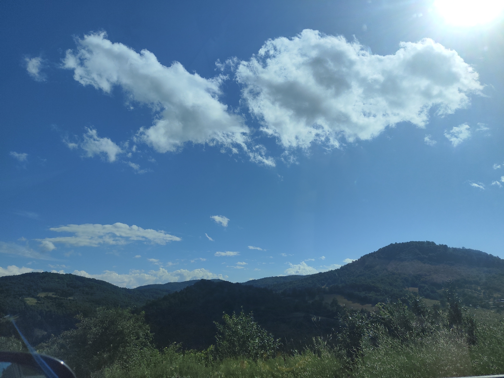 The image depicts a bright, sunny day with a clear blue sky. The upper portion of the image is dominated by a large, fluffy white cloud formation stretching horizontally across the frame. The sun is visible in the upper right corner, casting a bright glare and creating lens flare. Below the sky, a landscape of rolling, tree-covered hills is visible. The hills are a deep green, with some areas appearing darker due to shadows. The foreground consists of a grassy area with some trees and foliage, partially obscuring a car's side mirror and window on the lower left. The scene captures a natural, rural environment. Based on the provided context, this scene is located in Petrova, Romania.