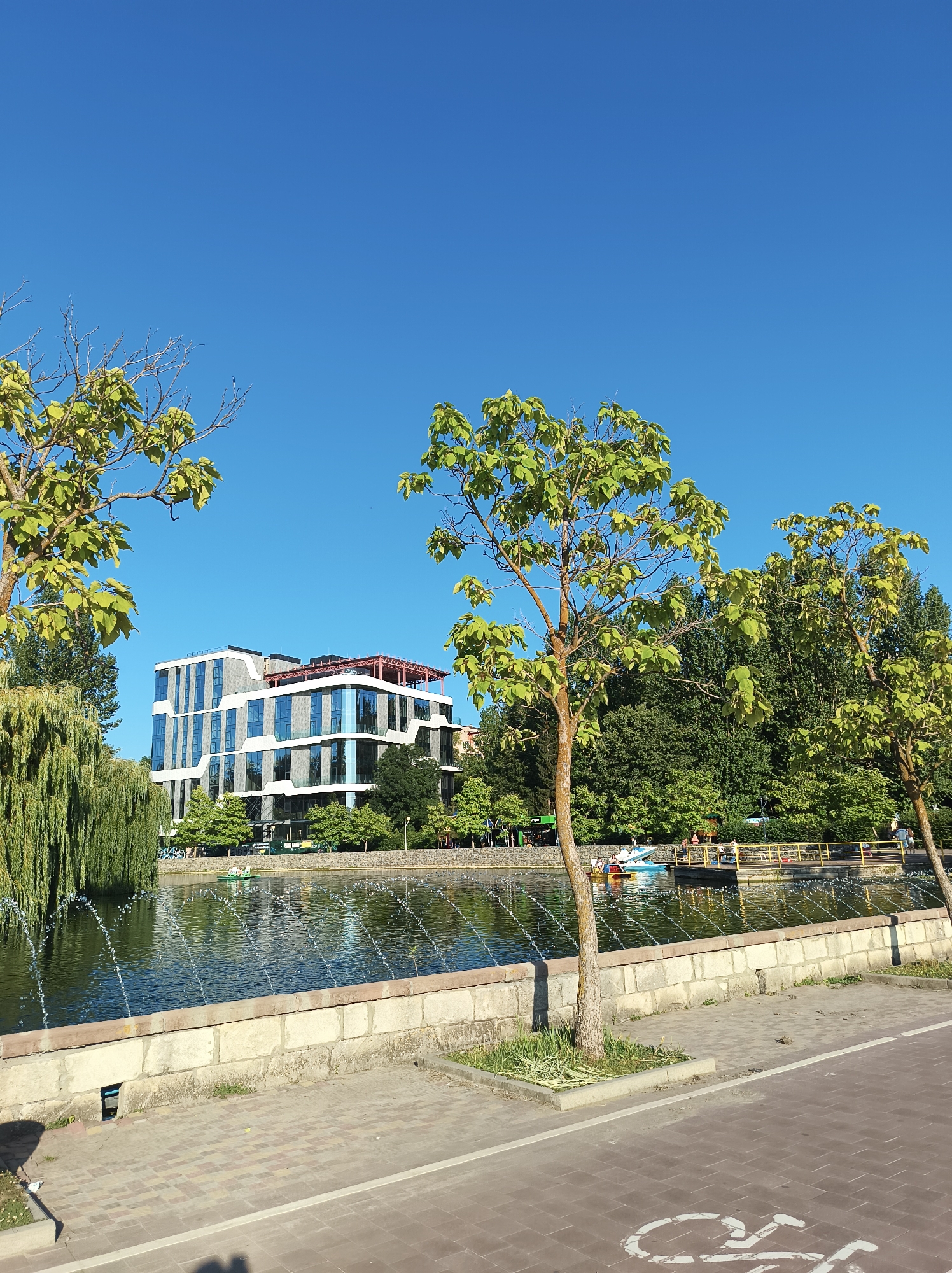 The image depicts a sunny day at a park in Ternopil, Ukraine. In the background, a modern, multi-story building with a distinctive facade featuring white geometric elements and a glass exterior stands against a clear blue sky. In front of the building, a body of water, likely a pond or lake, is visible. Several fountains are active, creating arcs of water.
The foreground shows a paved walkway with a white bicycle lane marking. To the left of the walkway, a low stone wall borders the water. Trees with green foliage are scattered throughout the scene; a weeping willow with long, drooping branches is prominent on the left, while two deciduous trees with lighter green leaves are positioned closer to the viewer.
On the water, a small pedal boat with several occupants can be seen. A yellow railing demarcates a walkway or pier extending over the water. In the distance, more trees and buildings are visible, suggesting an urban park setting. The overall impression is one of a pleasant recreational area.