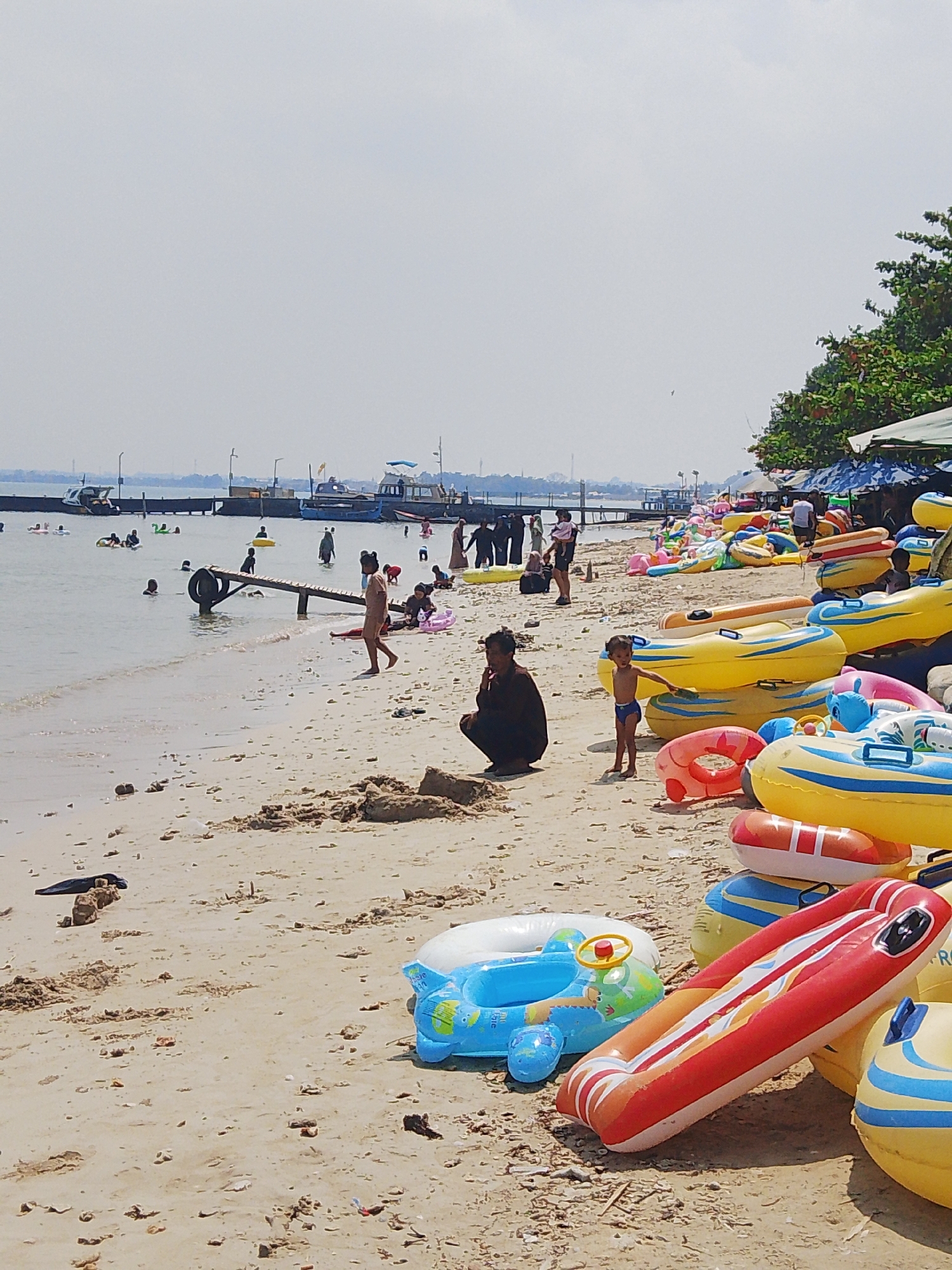 A group of people, including children and adults, are enjoying a day at the beach in Pejagan, Indonesia. The sandy shore is lined with various inflatable water toys, including tubes and ring floats in bright colors like yellow, red, and blue. Some of these inflatables are stacked on top of each other. Several people are in the shallow water, some swimming and others wading. A wooden pier extends into the water with boats docked alongside it. In the background, more people are visible further down the beach and near the water. The sky is overcast, and the overall scene depicts a lively beach atmosphere.