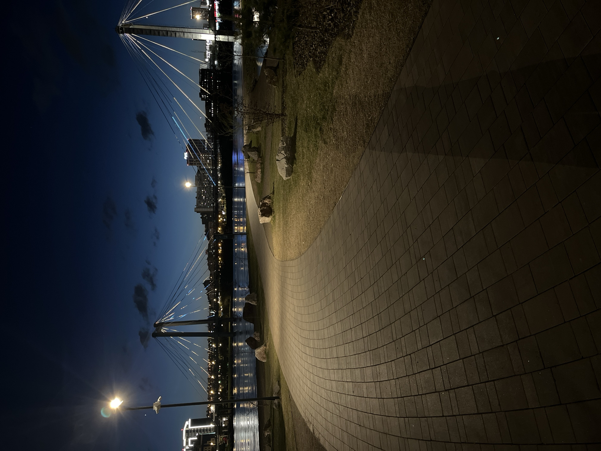 The image shows a nighttime scene featuring a curved brick pathway in the foreground, leading towards a body of water. A suspension bridge with lighted cables spans the water, with a city skyline visible in the background. The sky is dark blue, and streetlights illuminate the path and reflect on the water's surface. The edge of the path is lined with grass and a few rocks. The scene appears to be a city park or walkway near a river or bay.