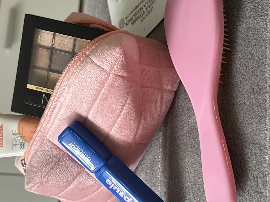 The image displays a collection of cosmetic and personal care items arranged on a grey, textured fabric surface in an indoor setting. The precise city and location within "Unknown, Unknown" are not identifiable from the visual evidence. A light pink, quilted, plush cosmetic bag, secured with a partially open zipper, occupies the central portion of the frame. Protruding from the bag's opening are a white tube labeled "OAVISAGE PRIME FACE BASE EXPERT" and a black rectangular eyeshadow palette containing nine neutral shades, marked "N Professional" and "FLN-Z". Resting on the grey surface in front of the bag is a blue tubular container identified as "Breez Tour Professional Capsule Lash Mascara". A pink hairbrush with yellow bristles extends into the lower right foreground. A white packaging sachet is partially visible on the right, displaying text that includes "LENGELAGE PULE VITAMIN ILLA MASK PACK". No individuals or active interactions are depicted. The background is softly blurred, indicating additional light-colored interior elements.