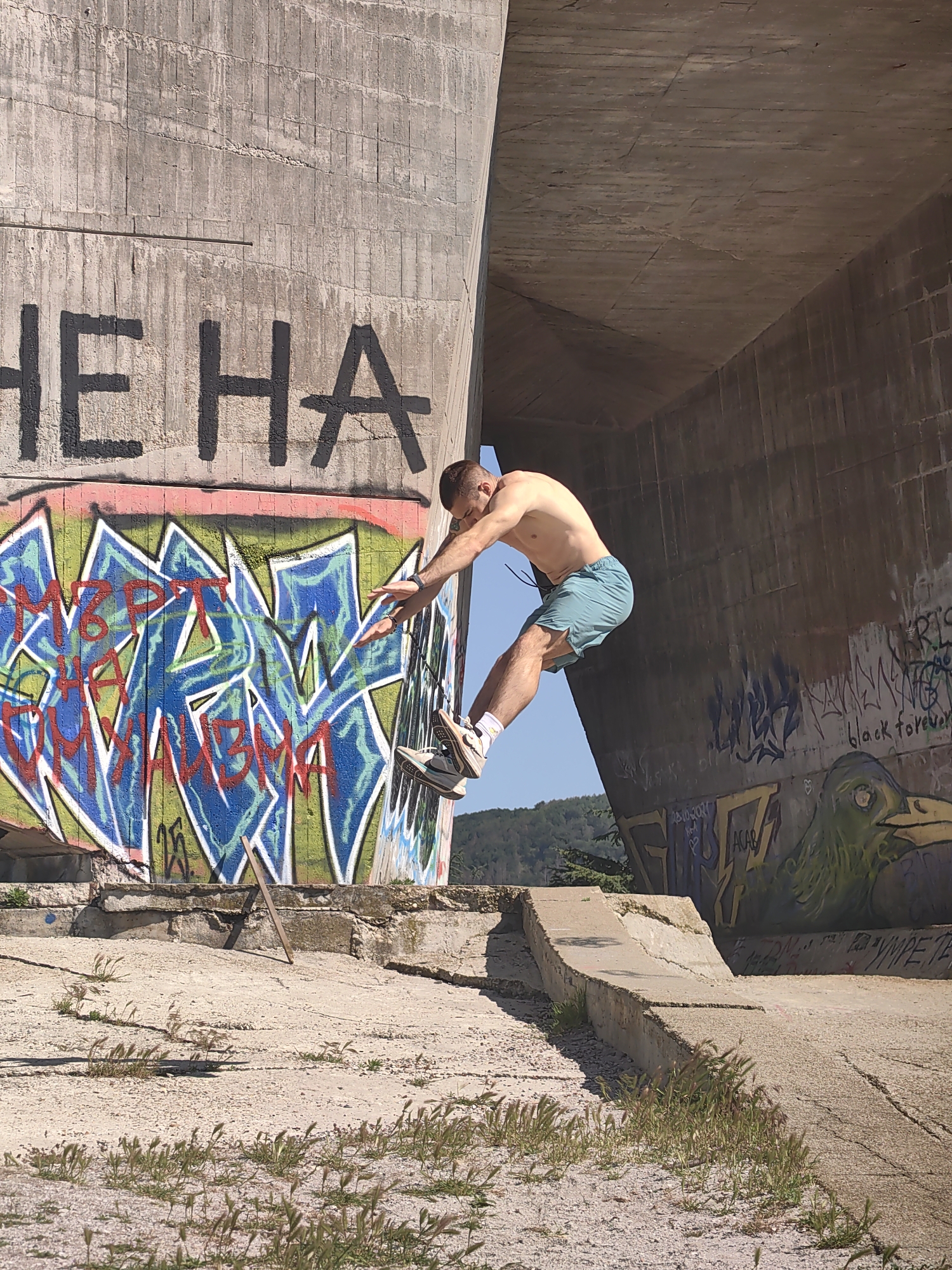 A man is hanging onto a concrete structure covered in graffiti in the city of Varna, Bulgaria. The structure appears to be part of a bridge or large building. The man is shirtless, wearing blue shorts, white socks, and sneakers. He is facing the wall, with his hands gripping the edge. The graffiti on the wall includes lettering in black and colorful designs. A grassy area and a hill with trees can be seen in the background.
