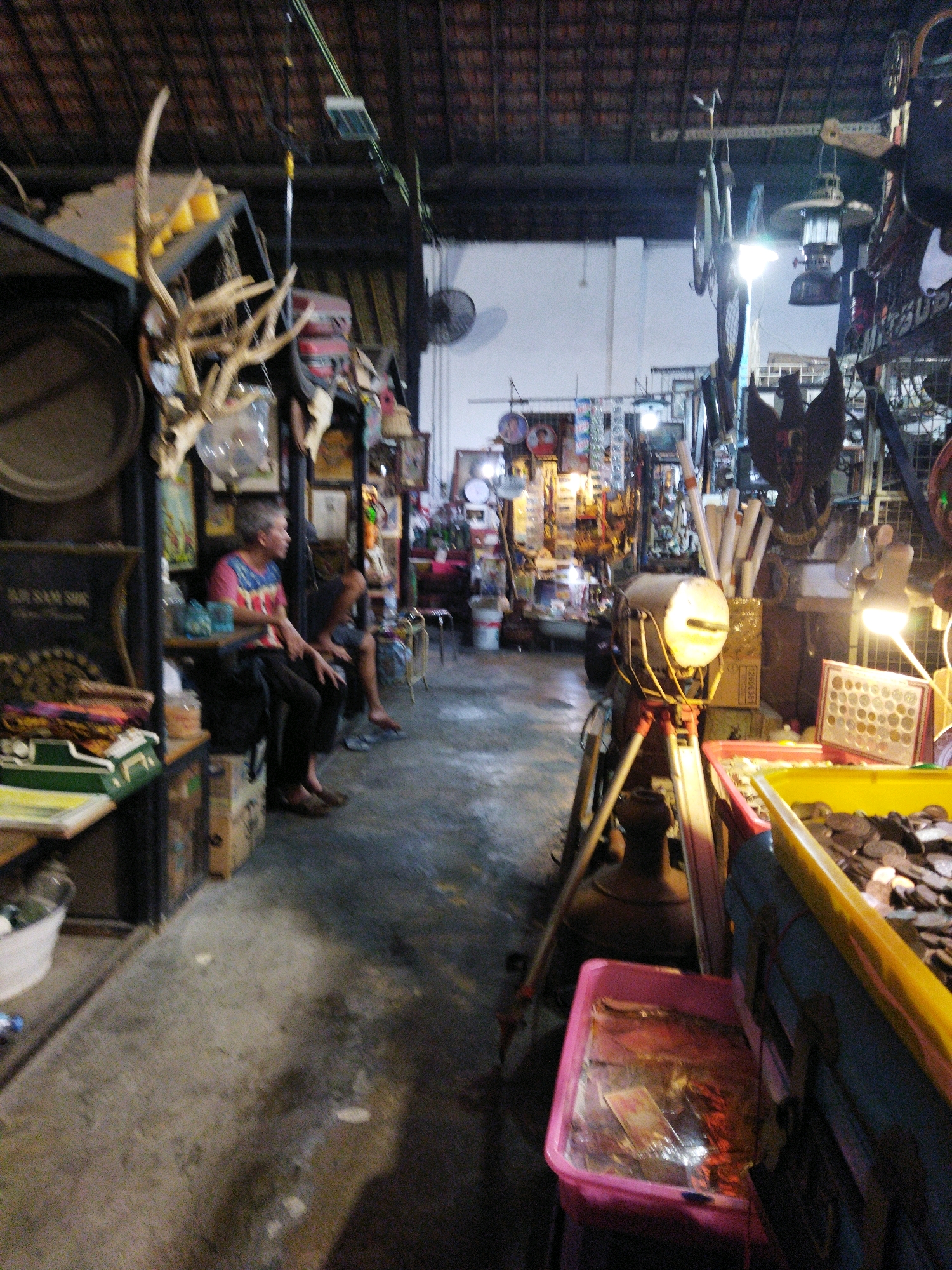 The image depicts an indoor market stall in Semarang, Indonesia, characterized by its eclectic collection of vintage items. The stall is dimly lit, with the ceiling constructed of dark, textured material, possibly tiles or wood. In the foreground, on the right side, a bright yellow plastic bin overflows with coins, suggesting a currency exchange or collector's stall. Adjacent to it, a pink plastic tub contains various items, including what appear to be old banknotes. A vintage spotlight on a tripod stands prominently near the bins.
Further back, two individuals are visible. A person wearing a colorful t-shirt and dark pants sits on a wooden crate, looking to their right. Another person is seated behind them, partially obscured. The background reveals shelves stacked with an assortment of objects, including antlers, framed pictures, and decorative items. Hanging from the ceiling are bicycles and other paraphernalia, contributing to the cluttered yet organized atmosphere of a curio shop or antique market. The floor is concrete, showing signs of wear and tear. The overall impression is that of a vibrant marketplace specializing in antique and second-hand goods.