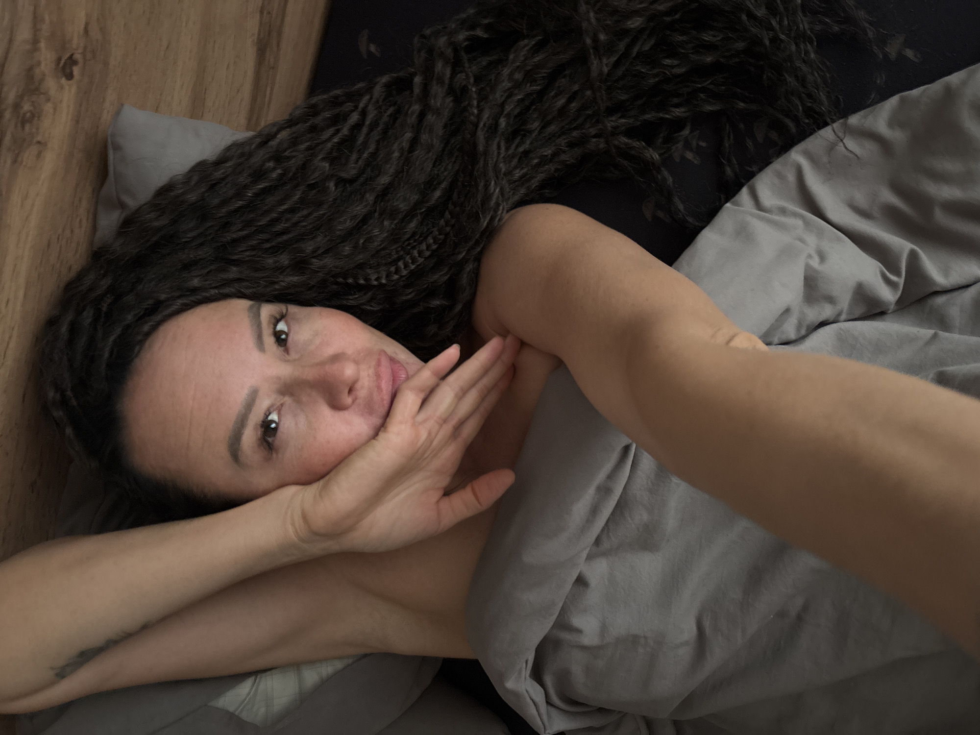 The image depicts a woman lying in a bed, viewed from an overhead perspective. She has long, dark, micro-braided hair fanned out across a dark grey fitted sheet on the right side of the frame. Her head rests on a light grey pillow, situated against a visible wooden headboard in the upper right quadrant. The woman has a light to medium skin tone. She is partially covered by a rumpled grey duvet, which extends from her mid-chest downwards, leaving her bare left shoulder and upper arm exposed. A faint, dark tattoo is visible on her left upper arm. Her right arm is bent, with her hand positioned near her chin and mouth, slightly obscuring the lower part of her face. Her gaze is directed towards the camera, and her lips are slightly parted in a subtle, neutral expression. The scene is an indoor bedroom setting with soft, ambient lighting.
