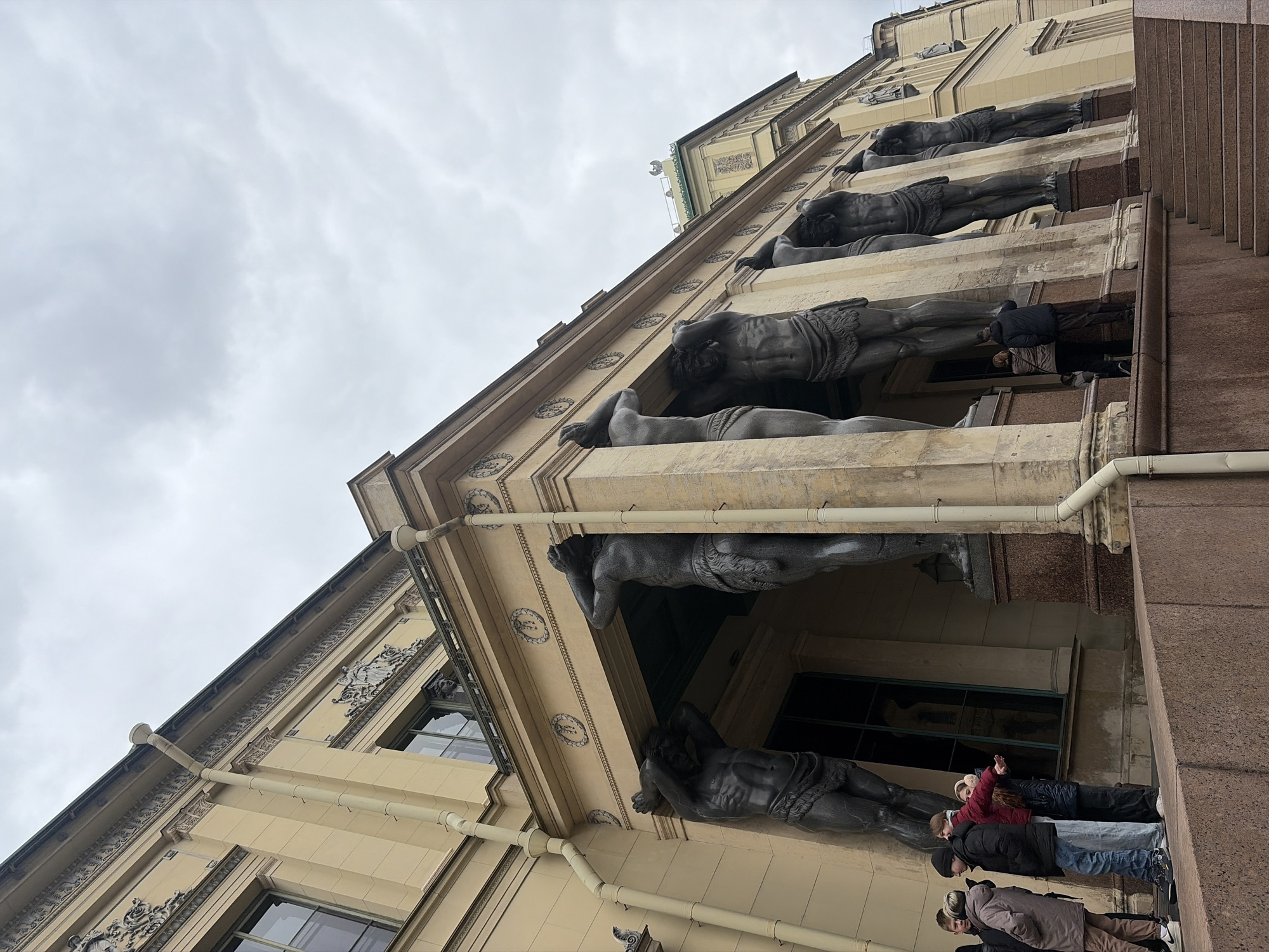 This image captures a low-angle perspective of a grand architectural facade, strongly suggesting the Atlantes of the New Hermitage Building in Saint Petersburg, Russia. The scene is illuminated by the soft, diffused light of an overcast day, with a pale grey, cloudy sky filling the background.
The building exhibits classical grandeur, featuring a light-colored, possibly stucco or stone, facade with intricate decorative moldings and cornices. The most prominent elements are a series of large, dark grey/black statues, likely Atlantes, depicted as powerful male figures supporting a projecting architectural feature above them. At least six of these muscular, semi-nude statues are visible in a row, each in a strained, crouched posture, appearing to bear the immense weight of the building on their shoulders and heads.
At the bottom right of the frame, a group of approximately five to six people stands on a paved area or steps adjacent to the building. Dressed in warm jackets and casual attire, some with hats, they are observed looking upwards, engrossed in the architectural details and the colossal statues. One person in a red jacket is seen gesturing towards the statues, possibly pointing something out to companions. A section of the building to the far right appears to be made of a reddish-brown, textured material, possibly granite, with vertical lines suggesting a structural element or stairs. No legible text is visible in the media file.