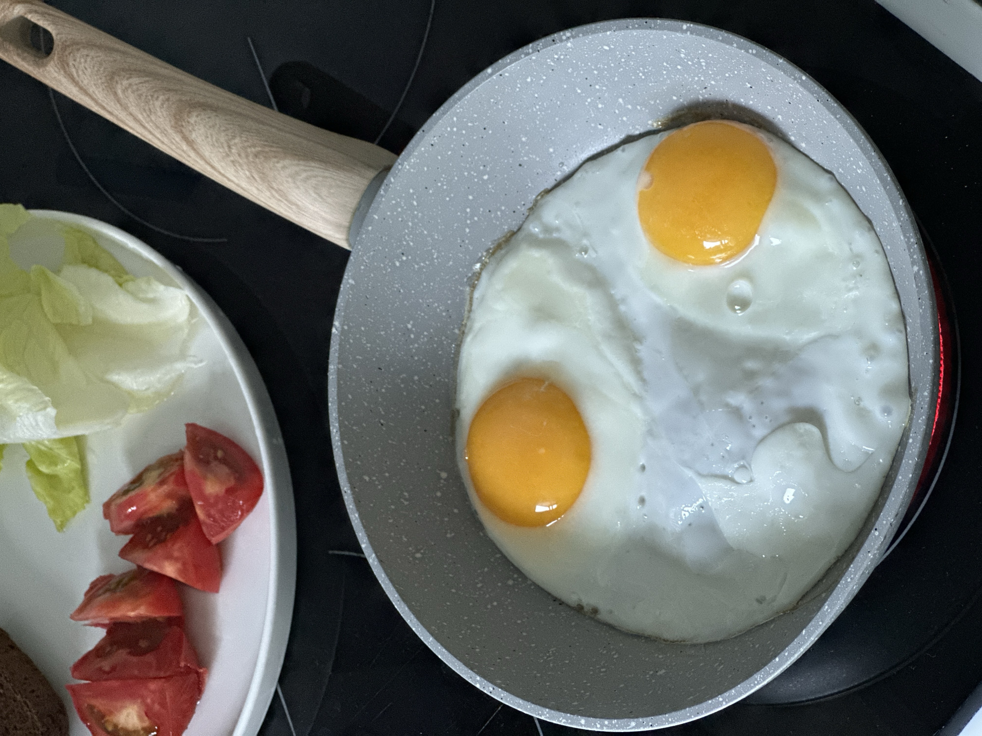 A close-up, high-angle shot captures two fried eggs with bright orange yolks sizzling in a speckled grey non-stick frying pan. The pan rests on a black stovetop, with a hint of red heat visible beneath it. To the upper left of the pan, a white plate holds a portion of chopped red tomatoes and some pale green lettuce leaves. The handle of the frying pan, made of light-colored wood with a grain pattern, extends diagonally upwards out of the frame. The image composition is tight, focusing on the food preparation. The scene appears to be in a kitchen, but no specific location in Moscow, Russia, can be determined from the visual information provided.