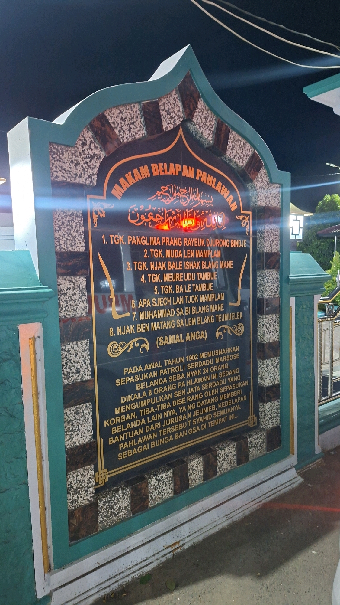 The image shows a monument commemorating "Makam Delapan Pahlawan" (Tomb of Eight Heroes) located in Lhokdagang, Indonesia. The monument is made of dark stone with gold and patterned inlays. Inscriptions in Indonesian are visible on the monument, including a list of eight names, a historical account, and the location "SAMALANGA". The monument is lit by artificial lights, and the background appears to be a dark, outdoor setting, likely at night, with some glimpses of greenery and structures. The monument itself has a distinctive arched top and is adorned with decorative borders.
