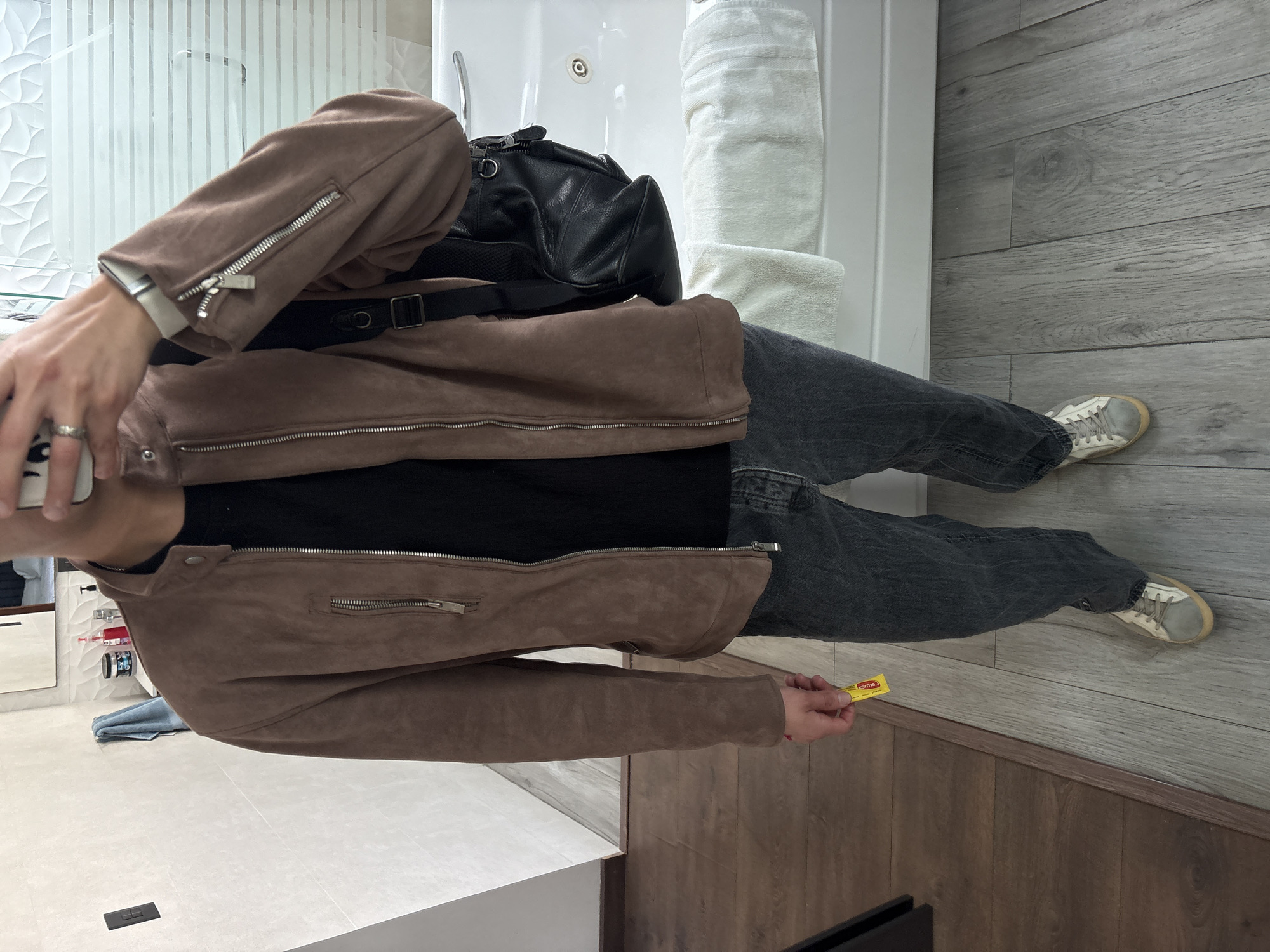 A person is standing in a bathroom with light gray wood-look flooring and white wall paneling. They are wearing a brown suede jacket, black t-shirt, dark gray jeans, and off-white sneakers. A black bag with a strap is slung over their shoulder. They are holding a small yellow packet in their right hand. The bathroom includes a white bathtub with jacuzzi jets and a stack of white towels. The image was taken from a first-person perspective, and a phone is visible in the upper left corner. The location, San Bernardino Tlaxcalancingo, Mexico, is not visually identifiable in the image.
