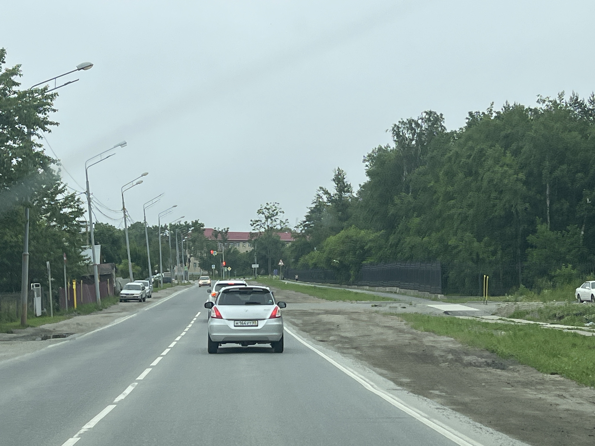 The image shows a road scene in Dolinsk, Russia, likely on a cloudy day. A silver Suzuki Swift is in the foreground, traveling down a paved road with a white dashed line. Several other vehicles, including a white sedan and a light-colored hatchback, are also visible on the road. On the left side of the road, there are trees, streetlights, and a fence. On the right side, there is a grassy area with a dark fence and more trees. In the background, buildings can be seen. The overall impression is of a suburban or semi-rural road.