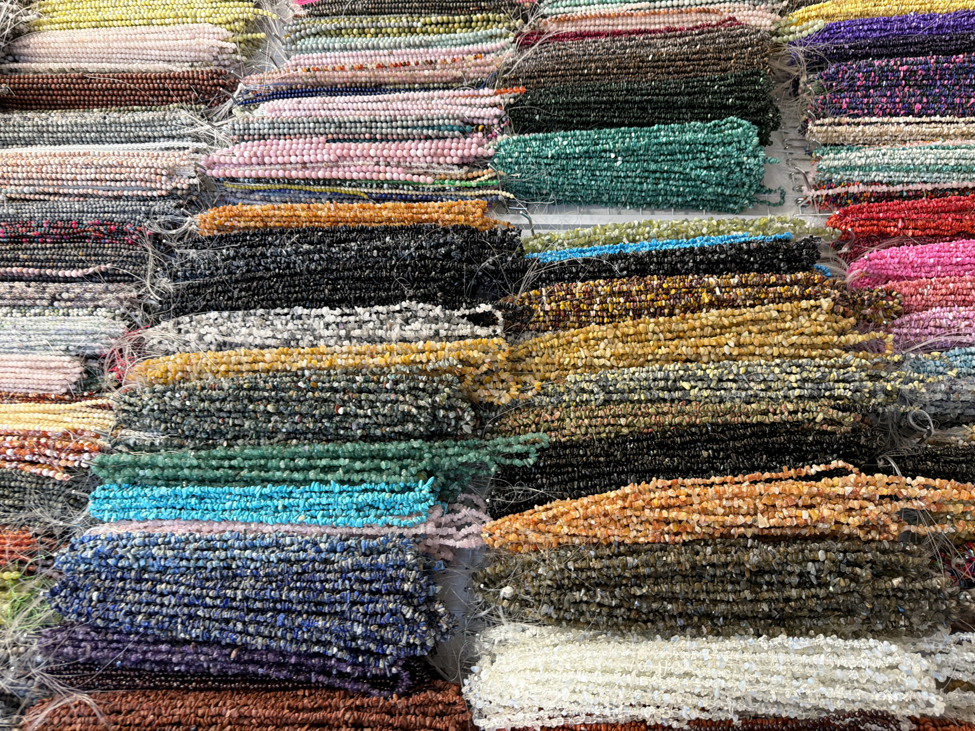 This media file showcases a vibrant and diverse collection of gemstone beads, meticulously arranged in stacked rows. The beads, likely intended for jewelry making, vary greatly in color, size, and material, creating a visually rich tapestry.
The subjects are primarily objects – specifically, numerous strands of semi-precious gemstone chips. These are presented in a flat lay or organized display, filling the frame from edge to edge. There are no people visible in the image, nor is any activity or event taking place beyond the static presentation of the merchandise.
Notable details include the sheer volume and variety of the beads. Colors range from pale yellows and pinks to deep greens, blues, purples, blacks, and fiery oranges and reds. Some strands feature a uniform color, while others are a mix of hues within a single strand. The texture of the beads appears to be that of small, irregular chips, giving them a natural, unpolished look. Fine white threads or strings are visible, holding the strands together or keeping them organized. The lighting suggests an indoor setting, possibly a market stall or a shop, with no obvious indications of time of day or weather. The context provided suggests this scene is located in Kuta, Indonesia, likely a place where such artisanal goods are sold. There is no visible text within the image.