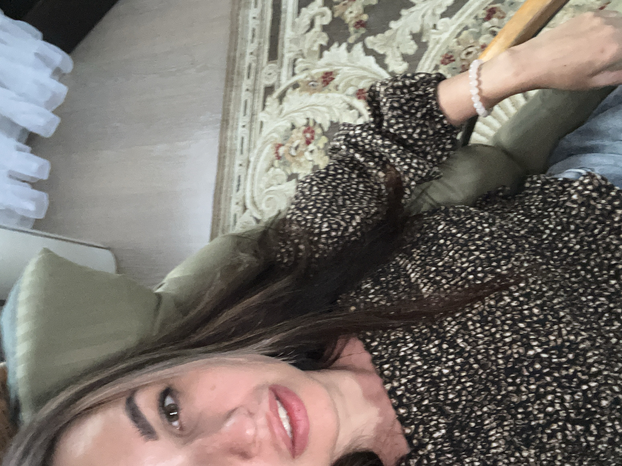 A medium shot shows a woman sitting in a chair, with her face close to the camera. She has long, dark brown hair with blonde highlights, and her eyes are a light brown. She is wearing a dark-colored blouse with a white speckled pattern, and a light-colored beaded bracelet on her right wrist. The background shows a beige carpet with a floral pattern, a light-colored wall, and what appears to be white drapery.