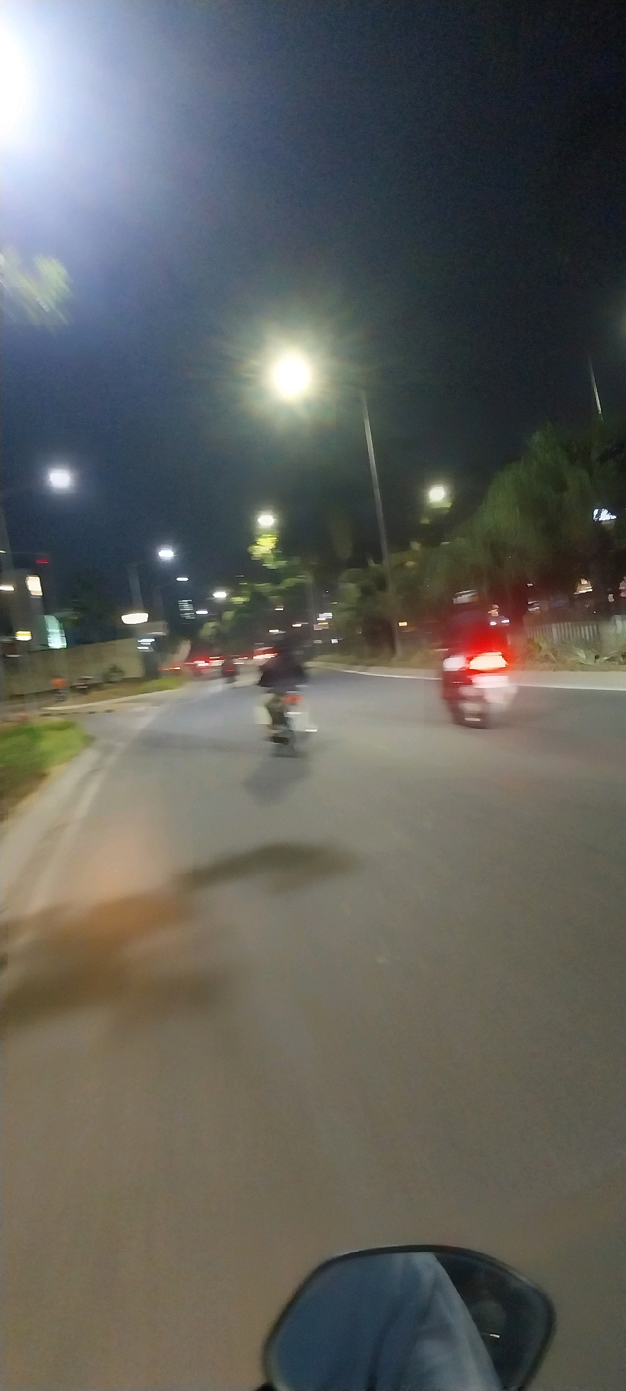 The image captures a night scene on a road in Medan, Indonesia. The perspective is from a moving vehicle, possibly a motorcycle, looking forward. Streetlights illuminate the road, with one particularly bright light in the upper left quadrant of the image. Several motorcycles are visible on the road, some in motion, with blurred figures of riders. One motorcycle in the foreground, on the right, has its red taillight illuminated, indicating it is also moving. Trees and buildings line the sides of the road, partially obscured by the darkness and motion blur. The overall atmosphere suggests a typical urban night on a street in Medan.