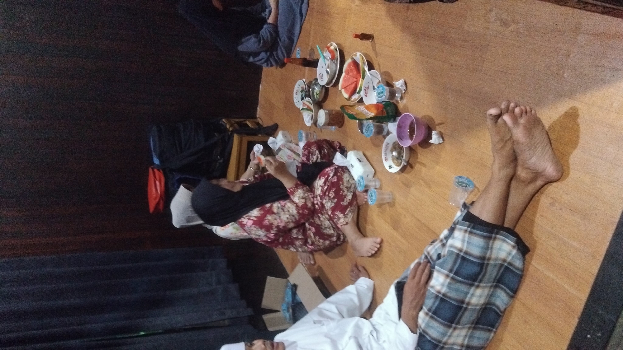 A group of people are seated on a wooden floor, in what appears to be a dining area. A man in a white shirt and a sarong is in the foreground. To his right, a woman wearing a patterned red dress and a black hijab is eating. To the right of the woman, another person wearing a blue hijab is seated. Several plates of food, bowls, drinks, and tissues are spread on the floor. The background shows a dark wall and curtains.