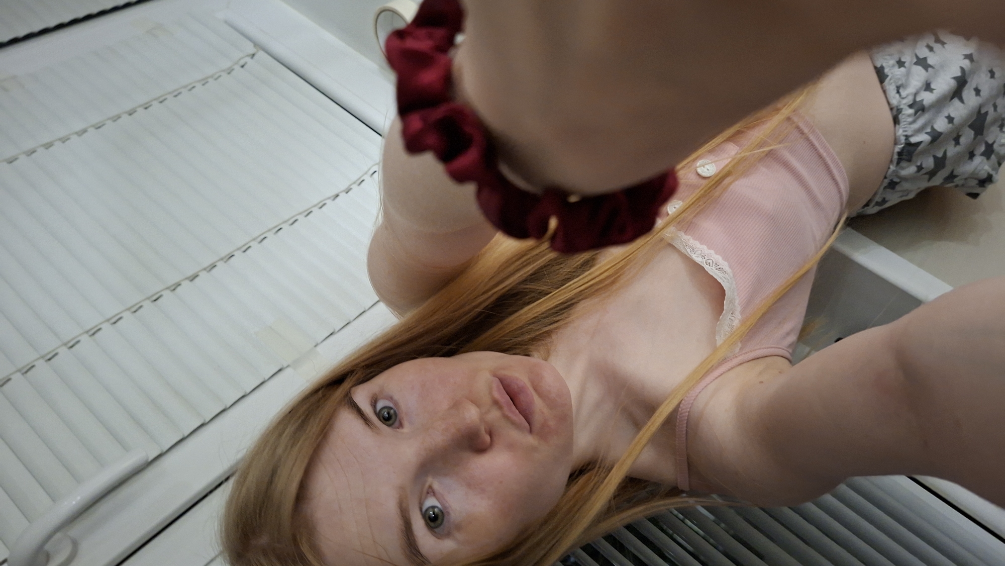 The image shows a woman with long, reddish-blonde hair taking a selfie. She is wearing a pink tank top with white lace trim and grey shorts with white stars. A red scrunchie is on her wrist. The background includes white blinds and what appears to be a bathroom sink. There is no definitive evidence to confirm the location is Chaikovsky, Russia.
