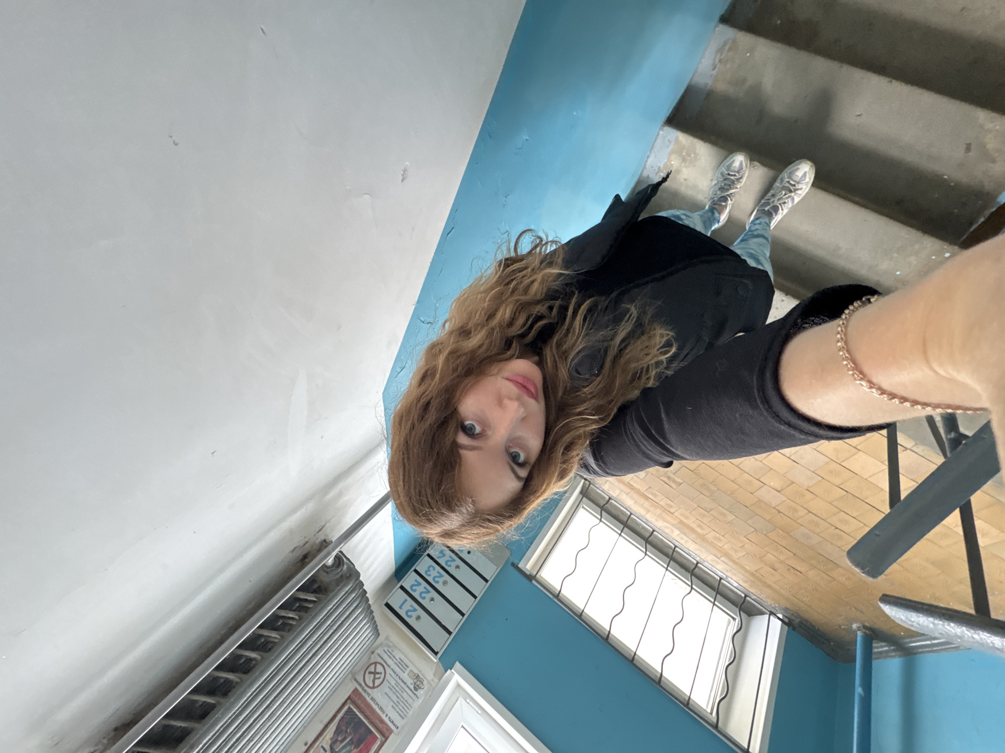 A selfie captures a young woman in a stairwell or common area of what appears to be an apartment building in Chaikovsky, Russia. The image is taken from a slightly upward angle, with the woman looking directly into the camera. She has long, wavy, light brown hair and blue eyes, wearing a black top or jacket, and a golden chain bracelet on her right wrist, which is visible in the foreground as she holds the camera.
The setting features a prominent white wall on the left and a vibrant light blue wall separating different levels. Concrete stairs are visible in the upper right, leading upwards. On the lower left, beneath the white wall, is a silver-colored metal radiator. Above the radiator, on the white wall, there's a sign with a no-smoking symbol and Cyrillic text, which is partially obscured but confirms a Russian-speaking environment.
Further right, mounted on the blue wall, is a unit displaying numbers "21", "22", "23", and "24" stacked vertically, likely indicating apartment numbers or mailboxes. Below these numbers, two windows with security bars are visible, allowing natural daylight into the space. The floor below the windows is covered with light brown or beige checkerboard-patterned tiles. The woman is wearing light-colored, possibly silver or white, athletic shoes with visible laces. The overall impression is that of an older, utilitarian residential building interior.
