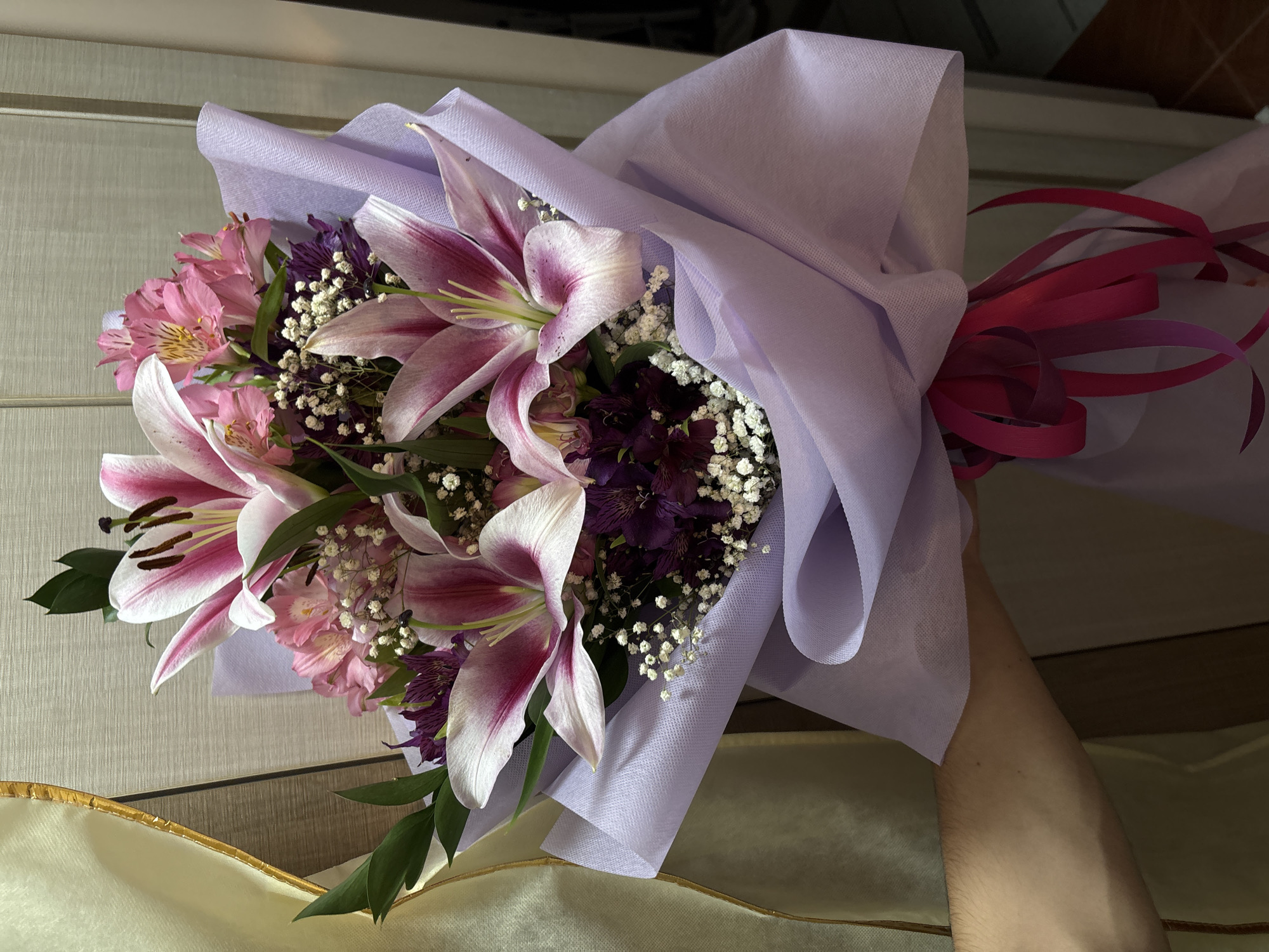A vibrant bouquet of fresh flowers is prominently displayed, held by a person's forearm. The bouquet primarily features large, striking pink and white lilies with visible dark brown stamens. Complementing these are clusters of smaller pink alstroemeria flowers, delicate white baby's breath (Gypsophila), and deeper purple accent flowers, possibly statice.
The flowers are elegantly wrapped in a light lavender or pastel purple non-woven fabric, which is gathered and tied at the base. A bright fuchsia or reddish-pink ribbon secures the wrapping, with several curled strands adding a decorative touch. The person's arm, visible in the lower right, has a natural skin tone and fine hair, holding the bouquet upwards.
The background consists of light-colored, horizontally textured panels or blinds in the upper portion of the image, suggesting an indoor setting. In the lower left, a cream-colored fabric with a golden trim is visible, likely part of furniture such as a cushion or armrest. There is no visible text in the image. While the location context of Burgas, Bulgaria is provided, no specific visual cues within the image confirm this location. The overall scene depicts a beautiful floral arrangement, likely presented as a gift or for display.
