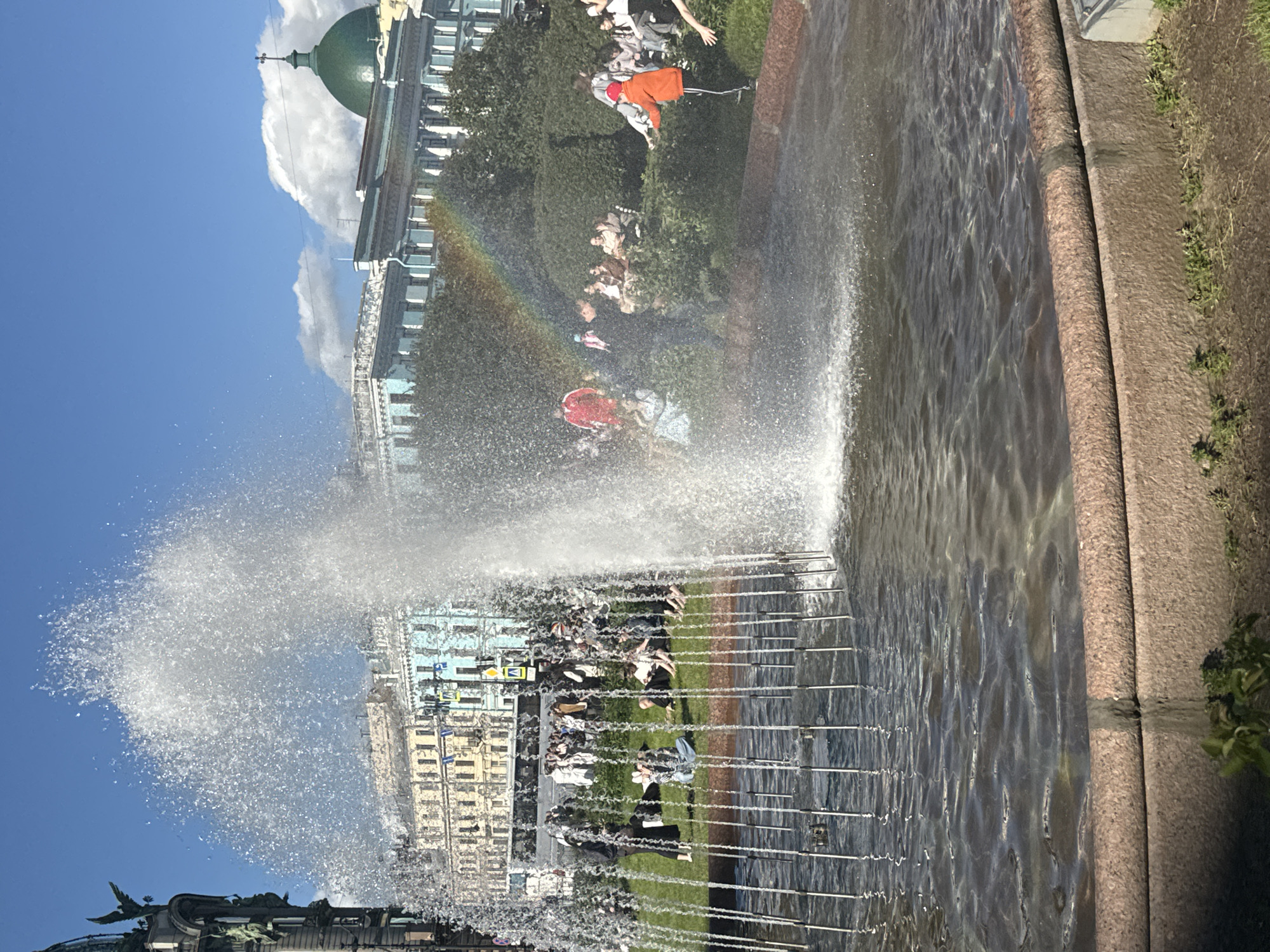 This is a photograph of a fountain in Palace Square, Saint Petersburg, Russia. The fountain is in the foreground with water spraying upwards. In the background, there are several historic buildings, including the General Staff Building and the Winter Palace. A rainbow is visible in the mist from the fountain. Several people are sitting on the grass around the fountain, enjoying the sunny day. The sky is blue with some white clouds.