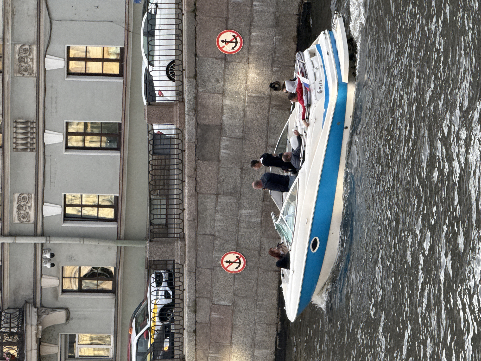 The image captures a scene along a canal in Saint Petersburg, Russia, with a building on the left and a boat on the right. The building features multiple windows with yellow-tinted glass, some of which appear to have reflections of the sky. Decorative architectural elements are visible on the facade. A white car is parked near the canal bank, partially obscured by railings.
The boat, a white vessel with a blue stripe, is moving through the water, creating ripples. Several people are on board, appearing to be tourists on a sightseeing tour. One person is wearing a red jacket, and another is holding a phone. A flag, possibly Russian, is attached to the boat.
A circular sign with a red border and a black anchor crossed out in red is affixed to the stone wall along the canal, indicating a prohibition against anchoring. The water is choppy, suggesting it might be a breezy day. The overall impression is one of urban activity and leisurely canal travel. The lighting suggests it is daytime, possibly late afternoon or early evening, given the warm glow in the windows. The architecture and the presence of a canal are strong indicators of Saint Petersburg. Text is visible on the boat, appearing to read "ВЕГА" (VEGA).