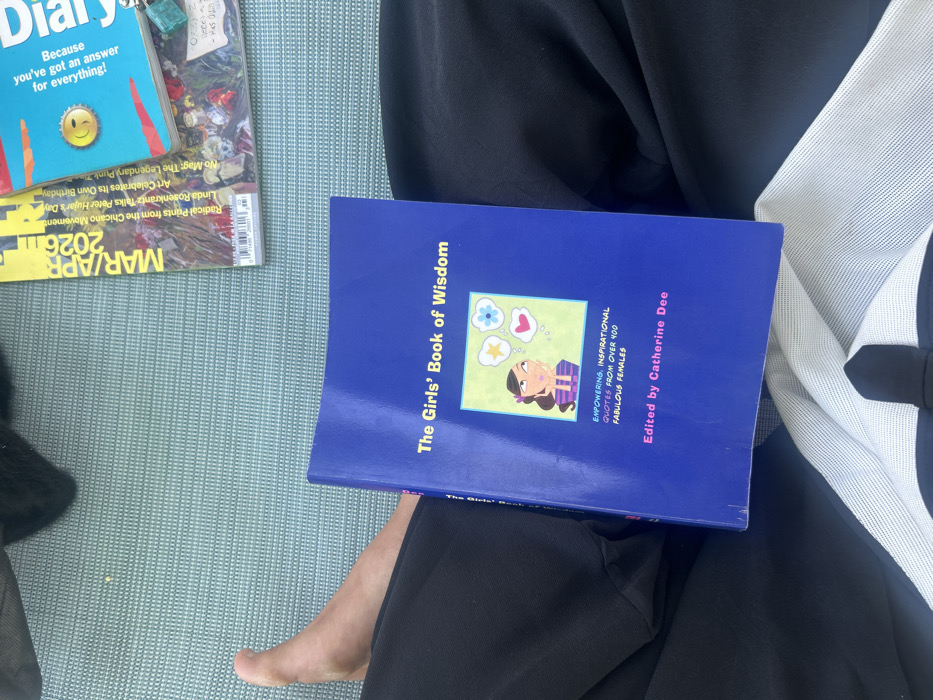 The image depicts an overhead view of a person seated outdoors in Los Angeles, United States of America. The subject's lower torso, legs, and a bare left foot are visible. They are wearing dark-colored trousers or shorts and a white garment underneath. A blue hardcover book titled "The Girls' Book of Wisdom," edited by Catherine Dee, rests on their lap. The book cover features a cartoon illustration of a girl with thought bubbles containing a star, a heart, and flowers, and includes the subtitle "Empowering, Inspirational Quotes From Over 400 Fabulous Females." To the upper right, two magazines are stacked on a light blue, textured outdoor surface. The topmost magazine has a blue cover with a smiley face and text partially visible, including "Because you've got an answer for everything!" Beneath it, a magazine dated "MAR/APR 2026" is visible, displaying articles related to "LA Galleries," "Chicano Movement," "Peter Hujar," and "Punk Zine," confirming the Los Angeles context. A small, irregular teal-colored object rests on the top magazine. The scene is illuminated by natural light, indicative of an outdoor setting.