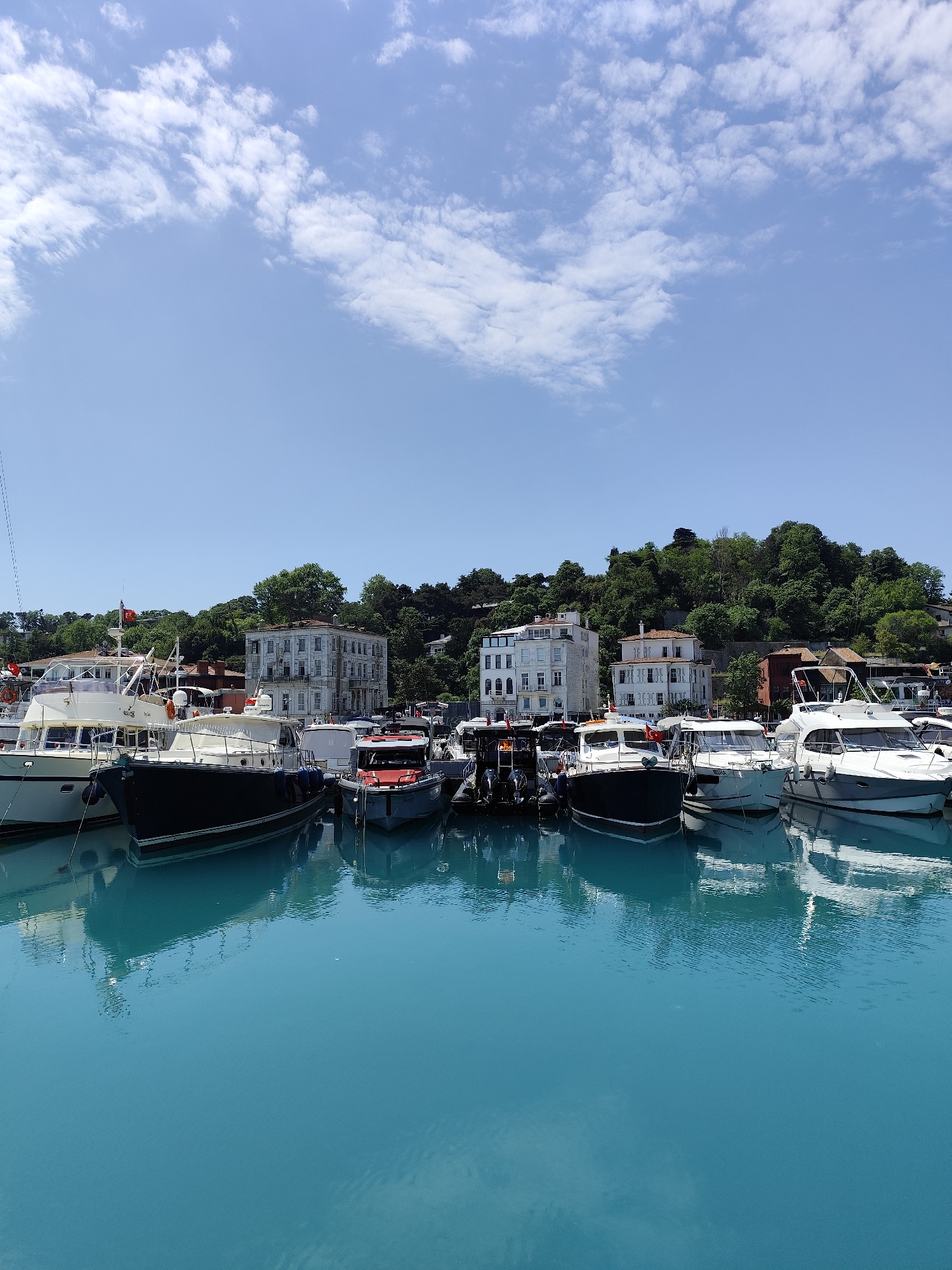 This vibrant image captures a sunny daytime scene at a marina in Sarıyer, Turkey. The sky, occupying the upper portion, is a clear, bright blue with scattered white cumulus clouds.
In the mid-ground, a lush, green hillside covered with dense trees forms the backdrop. Nestled among the foliage and along the waterfront are several elegant, multi-story buildings with classic architecture, predominantly white or light-colored, adding a sense of European charm to the landscape.
The foreground and central part of the image are dominated by a busy marina filled with numerous docked boats. These vessels are primarily recreational motor yachts and smaller cruisers, varying in size and color, with prominent white, dark blue, and grey hulls. They are tightly moored, suggesting a bustling harbor. A small red Turkish flag is visible on the mast of one of the boats on the left side of the frame, confirming the location context.
The water in the marina is exceptionally clear and a striking turquoise blue, reflecting the hulls of the boats and the overhead sky with remarkable clarity. Gentle ripples on the surface indicate calm conditions, enhancing the peaceful atmosphere of the scene. No discernible text is clearly visible on any of the boats or buildings.