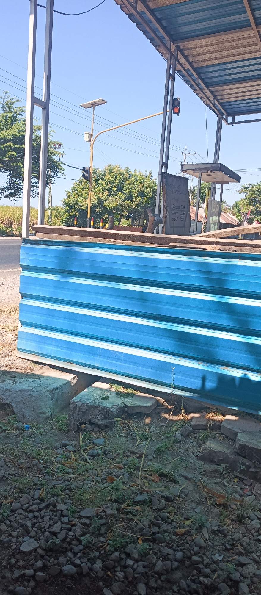 In Jatisari, Indonesia, a street scene is captured under a bright, clear blue sky. The foreground features a pile of blue corrugated metal sheets, stacked horizontally and supported by rough, gray concrete blocks. Beyond the metal sheets, a road is visible, bordered by dry grass and scattered small stones. In the middle ground, a traffic light hangs illuminated with a red light, indicating a stop. Behind the traffic light, trees with green foliage line the horizon, with some taller trees in the background. A solar panel is mounted on a pole to the left of the traffic light. A simple shelter with a corrugated metal roof is present on the right side of the frame, with a sign hanging from it. The overall impression is one of an outdoor, possibly industrial or construction site, setting adjacent to a road.