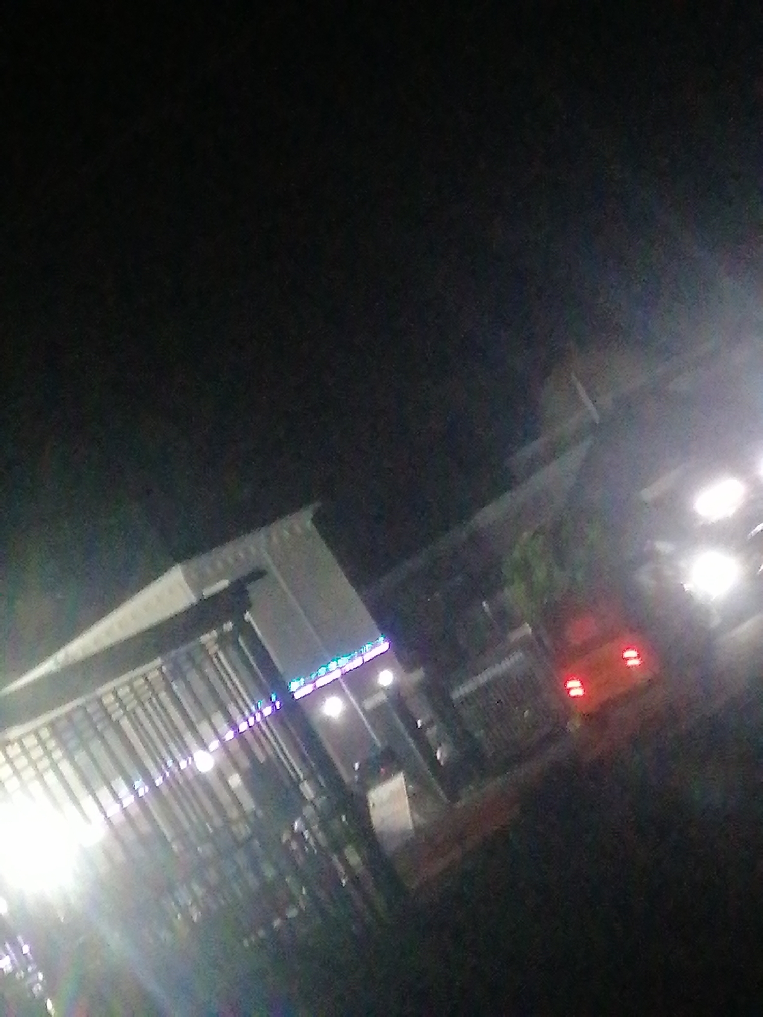 The image appears to be taken at night in an urban environment. A structure, possibly a building with an overhang or awning and some sort of metal bars, is visible on the left. Lights, including what might be decorative or LED strip lighting, are visible on the building. To the right, there are bright lights, most likely from vehicles, including at least one car with illuminated red taillights. The setting appears to be in an urban area. No identifiable landmarks or people are visible to confirm the location in Sabojibi, Nigeria.