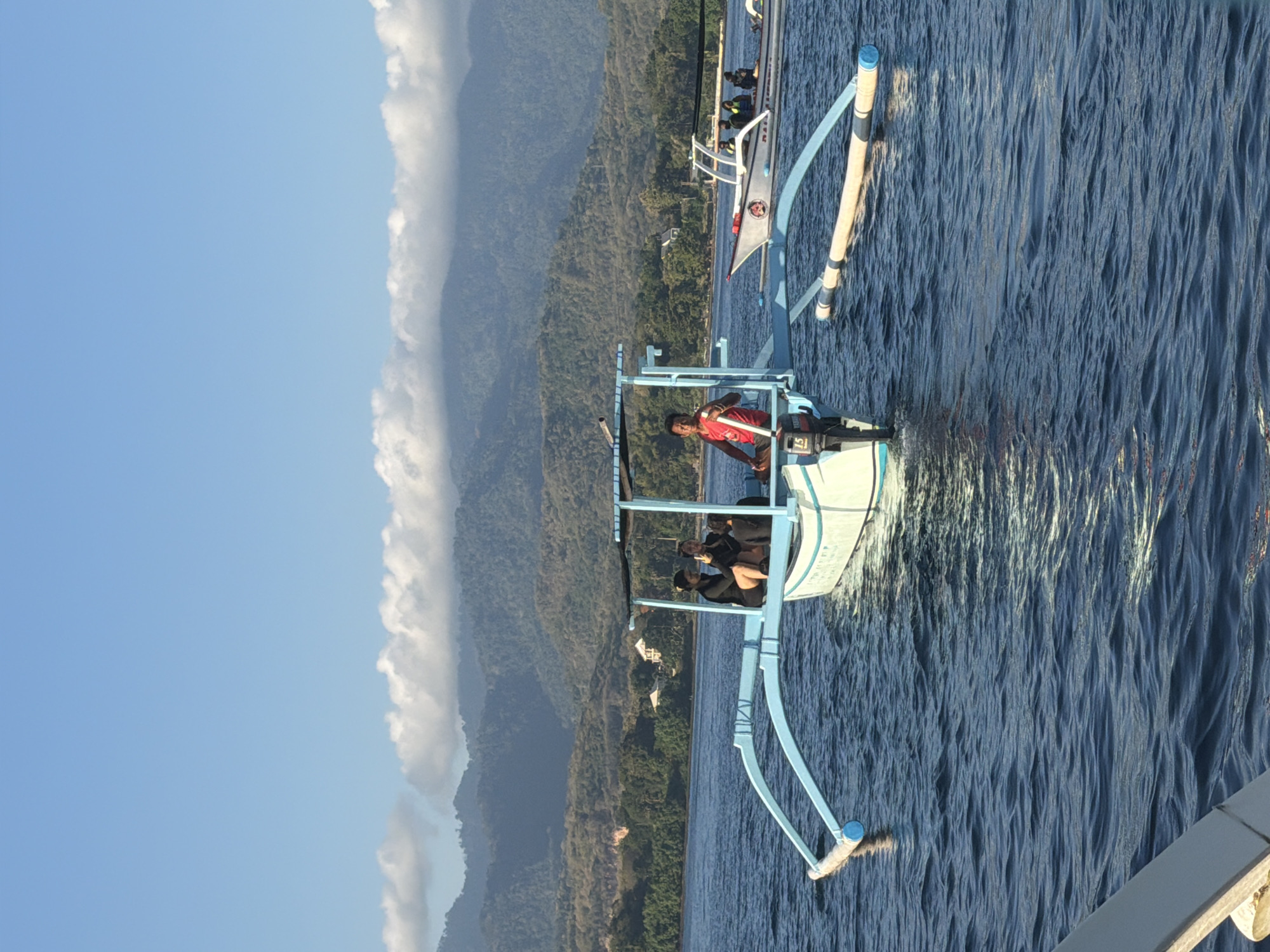 In the ocean of Unknown, Indonesia, a traditional outrigger boat with blue pontoons and a white hull carries three individuals. A man in a red shirt stands near the back, while two women in black attire sit towards the front, one of them holding a phone. The boat has a canopy for shade. In the background, another similar boat is visible, also with people on board. The water is calm with gentle ripples, and the sky is clear with a few scattered clouds. Lush green hills and mountains form the backdrop, with some buildings nestled among the trees on the shore. The scene appears to be a peaceful day on the water, possibly for a boat tour or island hopping.