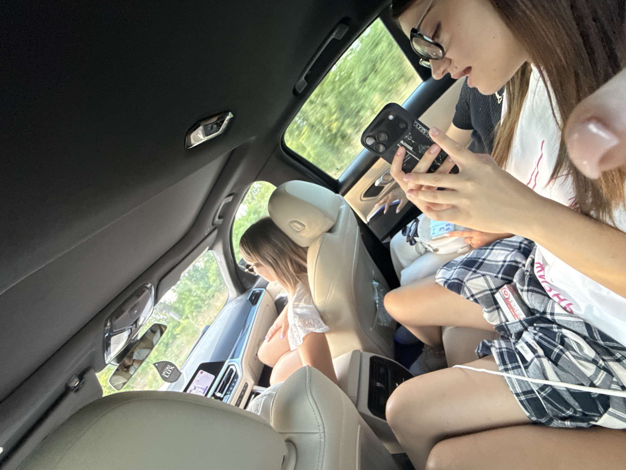 The image shows the interior of a car from a rear-facing perspective. Two young women are visible. The woman in the foreground on the right is wearing glasses and looking at her phone, which is held in her hands. She has long brown hair and is wearing a black top. The woman in the middle ground on the left is reclining and appears to be wearing a white top with lace trim and plaid shorts or a skirt. Her legs are extended. The car's interior features light-colored leather seats and a black headliner. A sun visor with a mirror is visible in the upper left, with a car air freshener hanging from it. The car is in motion, as indicated by the blurred green foliage visible through the windows, suggesting a rural or suburban setting. The scene is daylit, with sunlight entering the car.