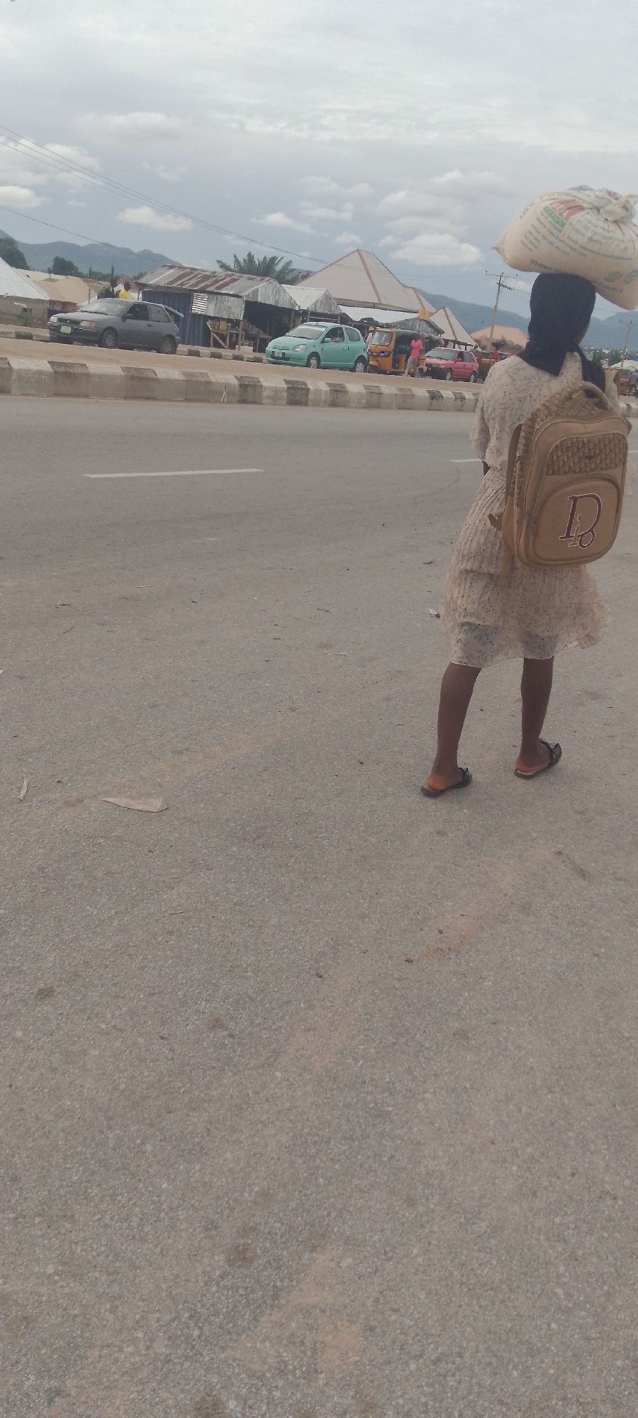 A young person wearing a light-colored dress and a backpack walks on a paved road. They have a large sack balanced on their head. In the background, there are buildings and several vehicles, including cars and a yellow tricycle. The sky is cloudy, and mountains are visible in the distance. The scene appears to be a street in Jalingo, Nigeria.