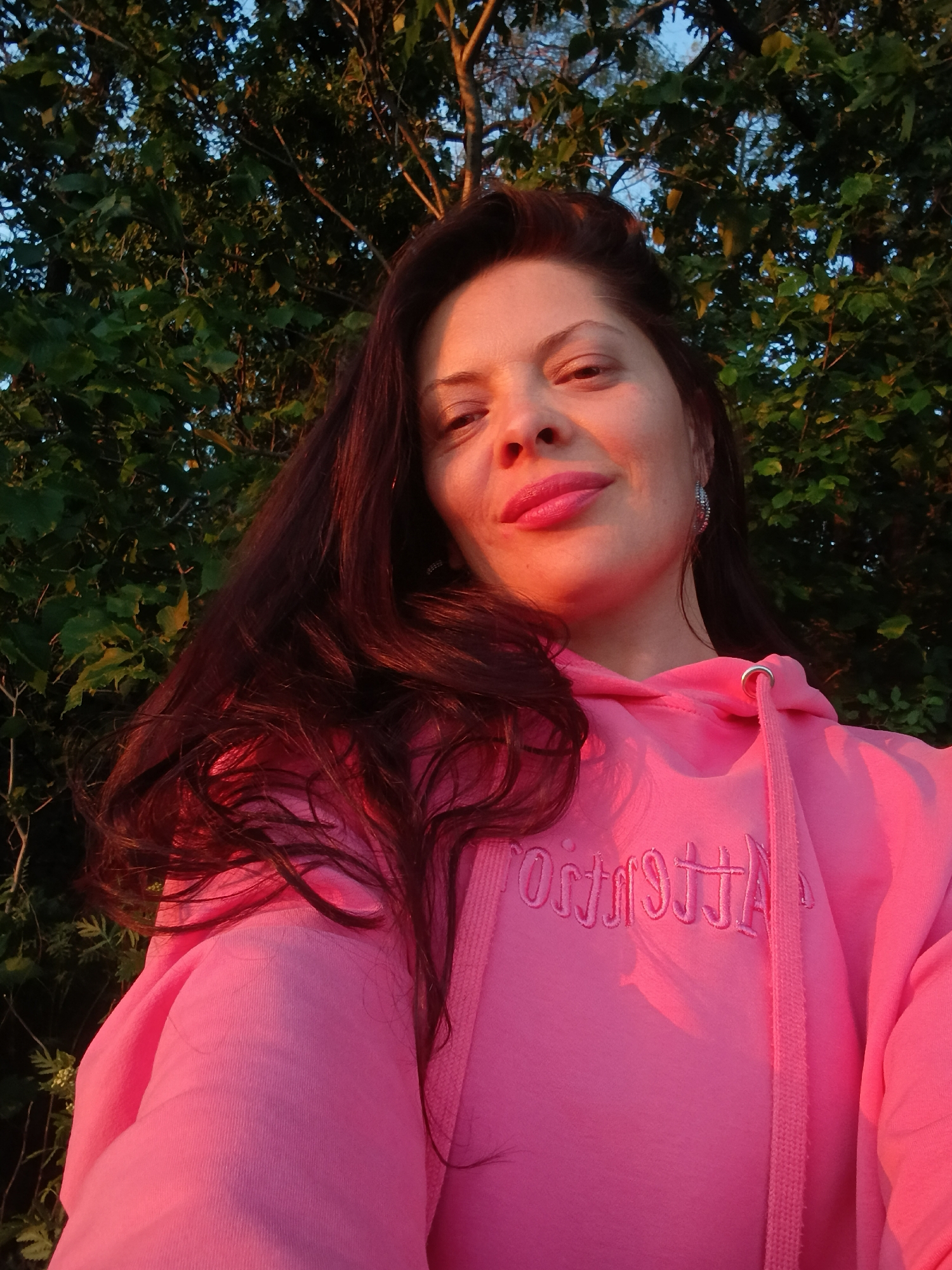 A woman with long, dark hair and pink lipstick is taking a selfie outdoors. She is wearing a pink hooded sweatshirt with embroidered text on the front. The background consists of lush green trees and a glimpse of the sky, suggesting a natural setting, possibly a park or garden within the city of Saratov, Russia. The lighting indicates it might be late afternoon or early evening, with the sun casting a warm glow on her face and clothing.