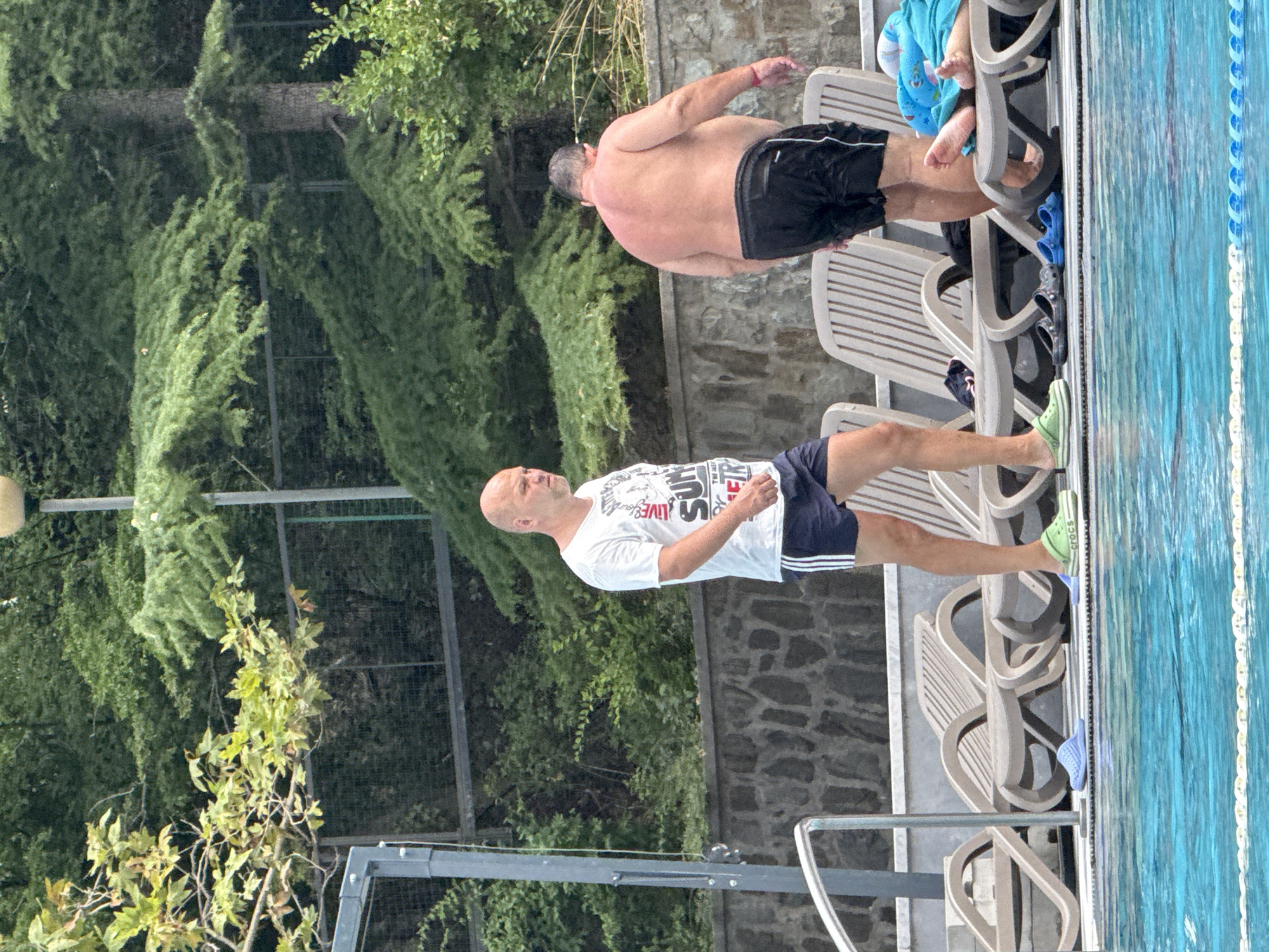 The image depicts an outdoor swimming pool area, likely in the city of Yalta, Ukraine, given the context. The focus is on two adult males. The man on the left is bald, wearing a white t-shirt with graphics and dark blue shorts with white stripes. He is walking towards the right side of the frame, looking slightly to his right. He is wearing green Crocs-style sandals. The man on the right is facing away from the camera, with his back to the viewer, and appears to be overweight. He is wearing black swim trunks. He is standing near several empty lounge chairs. The environment includes lush green trees and foliage in the background, a stone wall, and a metal fence. The swimming pool is in the foreground, with clear blue water and a white rope divider. There are several pairs of sandals and flip-flops scattered around the lounge chairs, suggesting recent activity. A blue towel is visible on one of the lounge chairs, with a person's feet peeking out from underneath it. The lighting suggests it is daytime, possibly overcast or late afternoon, as there are no harsh shadows.