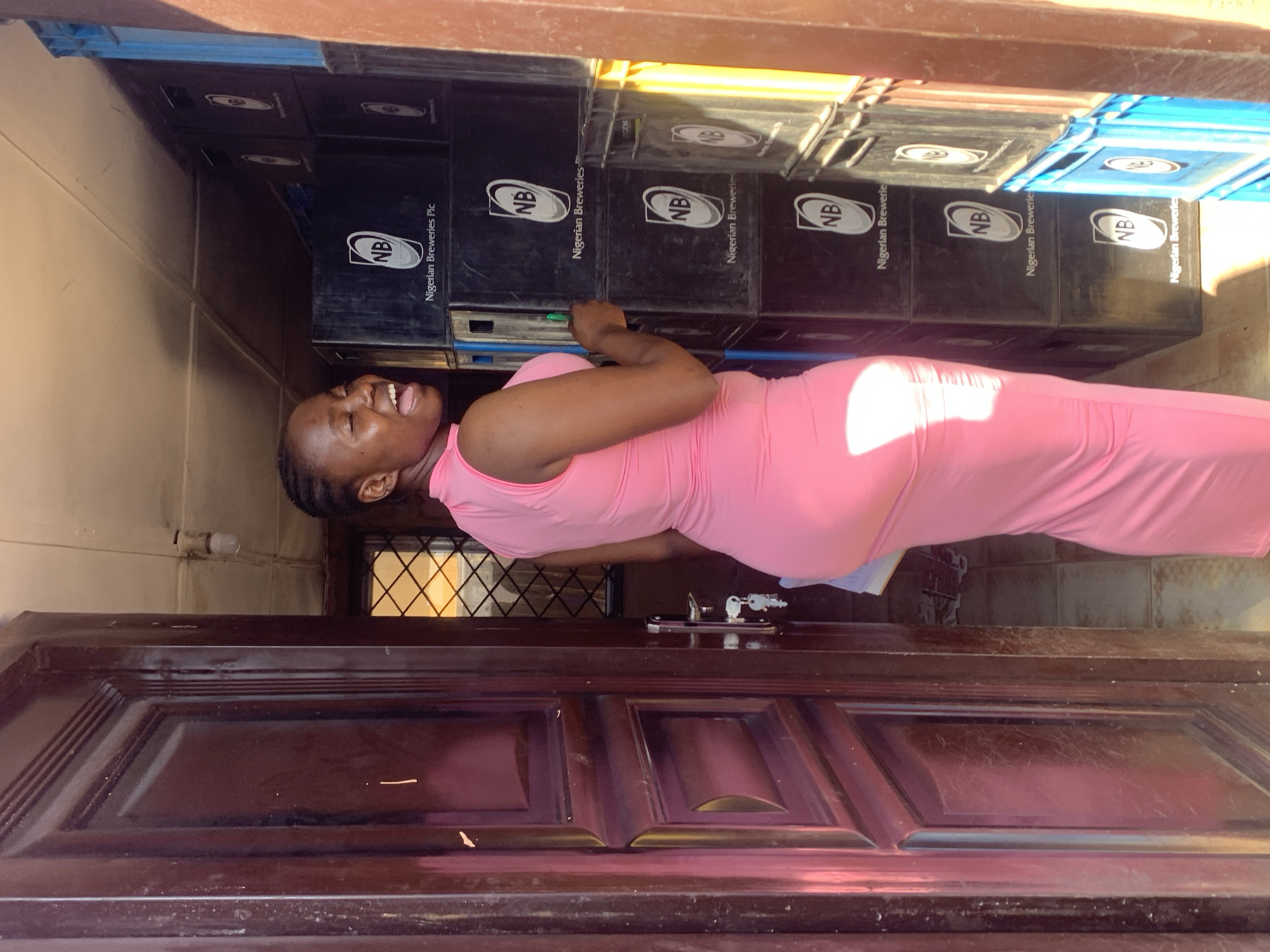The image shows a woman in a pink dress smiling and looking up at stacks of black crates labeled "NB Nigerian Breweries Plc." The crates are piled high, suggesting storage or delivery. A brown wooden door is positioned in the foreground, possibly indicating an entrance to a shop or storage area. The image's location is assumed to be Jos, Nigeria based on the brand on the crates.