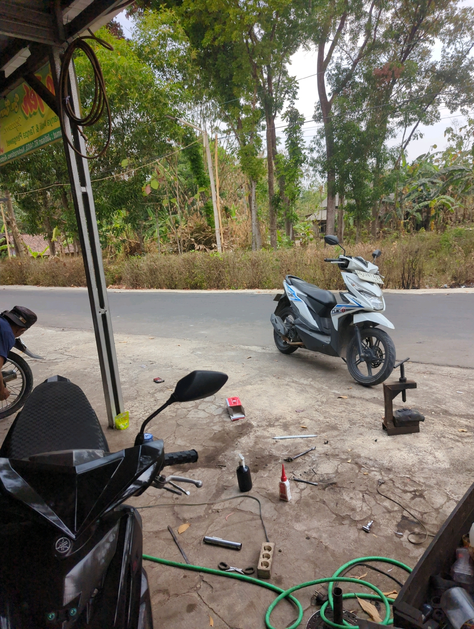This image depicts an outdoor scene at a roadside motorcycle repair stall in Kedungdowo, Indonesia. In the foreground, the front section of a black Yamaha motorcycle is visible, featuring a textured seat and a prominent Yamaha emblem on its front fairing. To the right of this motorcycle, a white Honda Beat scooter is parked, displaying a license plate that reads "AD 2287 GLO". The ground is a concrete surface littered with various tools and repair items, including a green hose coiled on the ground, a red-tipped bottle of adhesive, a spray bottle, a metal vise, a wrench, and what appears to be a small red toolbox or container. Behind the motorcycles, a gray paved road runs horizontally across the frame. A man wearing a dark blue shirt and a dark cap is partially visible on the left side of the image, attending to the wheel of a motorcycle that is mostly out of frame. The background is characterized by lush green trees and vegetation, with a few simple structures visible in the distance, suggesting a semi-rural or suburban setting. The overall atmosphere suggests an active, albeit somewhat cluttered, workspace.