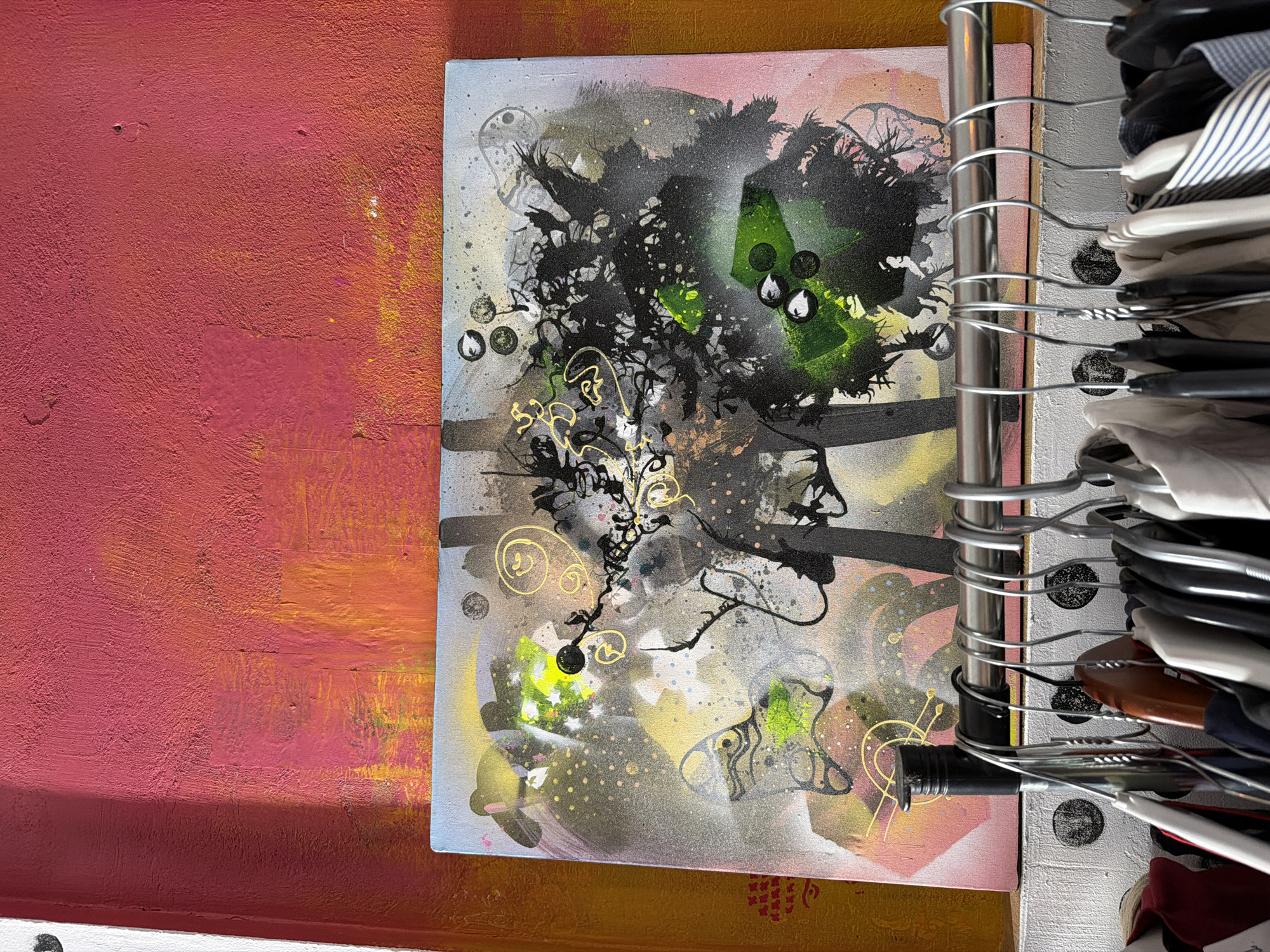 The image presents a close-up shot within a confined space, possibly a closet or storage area. A vividly painted wall, featuring a textured surface in varying shades of pink and orange, forms the backdrop on the left side of the frame. Positioned against this wall is a framed abstract artwork. The artwork showcases a chaotic composition of black, green, and gold splatters, lines, and amorphous shapes, resembling a stylized face in profile. To the right of the artwork, a vertical metal rod, likely part of a clothing rack, is visible, densely packed with clothes hangers and a variety of garments. The hangers create a layered effect, obscuring portions of the artwork and adding depth to the scene. The environment appears to be indoors. The image's location is undisclosed and cannot be ascertained from the image. Therefore, the specific location in Sofia, Bulgaria, is indeterminable.