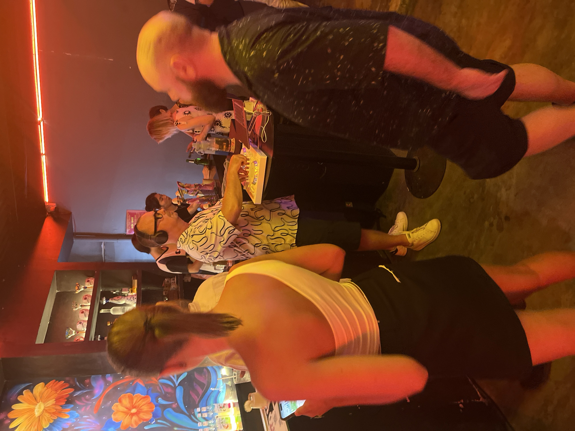 The image captures a bar scene, likely in Đà Nẵng, Vietnam. A mural featuring stylized flowers adorns the wall behind the bar counter. Several individuals stand and sit at the bar, engaged in various activities. One man with a beard leans over the counter, partially obscured by the camera's perspective. Another man wearing glasses and a patterned shirt interacts with a light-up panel. Patrons are seated at the bar and using cell phones. Bottles are arranged on shelves behind the counter. Artificial lighting illuminates the interior.