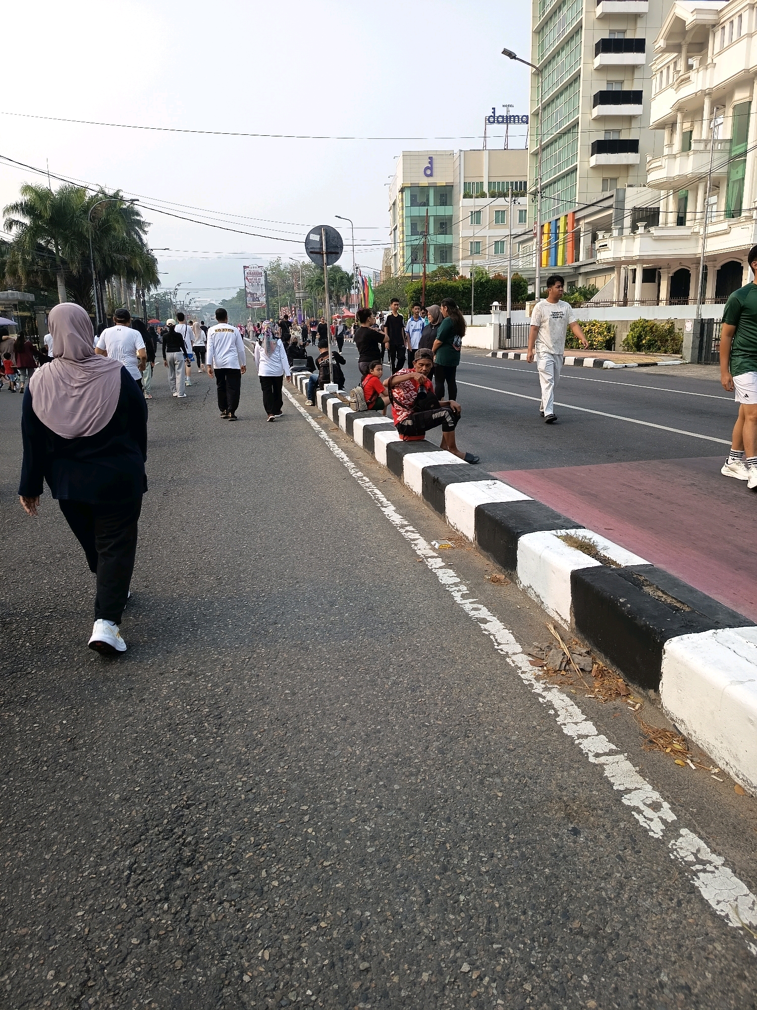 The image depicts a street scene in Padang, Indonesia, likely during a public event or morning exercise activity. In the foreground, a woman wearing a grey hijab and a dark outfit walks away from the camera on an asphalt road. Behind her, a group of people are walking in the same direction. They are dressed in casual sportswear, with many wearing white t-shirts. Some are seen walking alongside the road or on a designated path marked with red and white curbs. Buildings line the right side of the street, including a tall, modern hotel with the "daima" logo visible. Palm trees are present on the left side of the image. Several individuals are seen sitting on the curb, observing the activity. The overall atmosphere suggests a lively, outdoor gathering in a city environment.