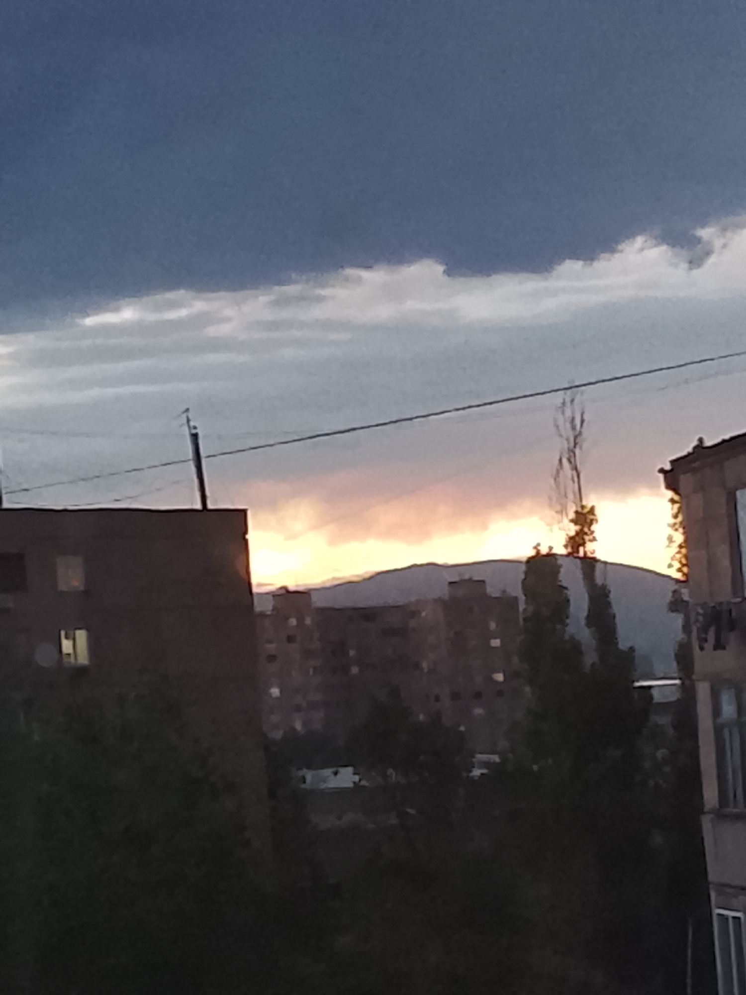 The image captures an urban or semi-urban landscape during either sunrise or sunset, characterized by a vibrant sky and silhouetted foreground elements. The upper portion of the frame features a dark blue-grey sky with a band of lighter white and grey clouds. Below this, a strong orange and yellow glow radiates from the horizon, indicating the position of the sun.
In the mid-ground, distant mountains appear as dark, hazy silhouettes against this bright light. Closer to the viewer, several multi-story buildings, likely residential apartment blocks, are visible. On the left, a large, dark building with a flat roof and a few rectangular, illuminated windows stands prominently. A utility pole with overhead wires extends across the scene, and a tiny bird is perched on one of these wires. To the right, another building's facade with windows is partially visible.
The lower half of the image is dominated by dark foliage from numerous trees, some appearing dense and others more sparse or bare. These trees, along with the buildings, are largely silhouetted against the brighter horizon, creating a stark contrast. In the bottom-left, a hint of light from a window is visible, and a faint white object, possibly a vehicle or structure, can be seen amidst the darkness in the lower mid-ground.
No discernible human activity or visible text is present. The scene conveys a tranquil and atmospheric mood, highlighting the transitional period of day and night. The architecture suggests a setting in a city or town with a mountainous backdrop.