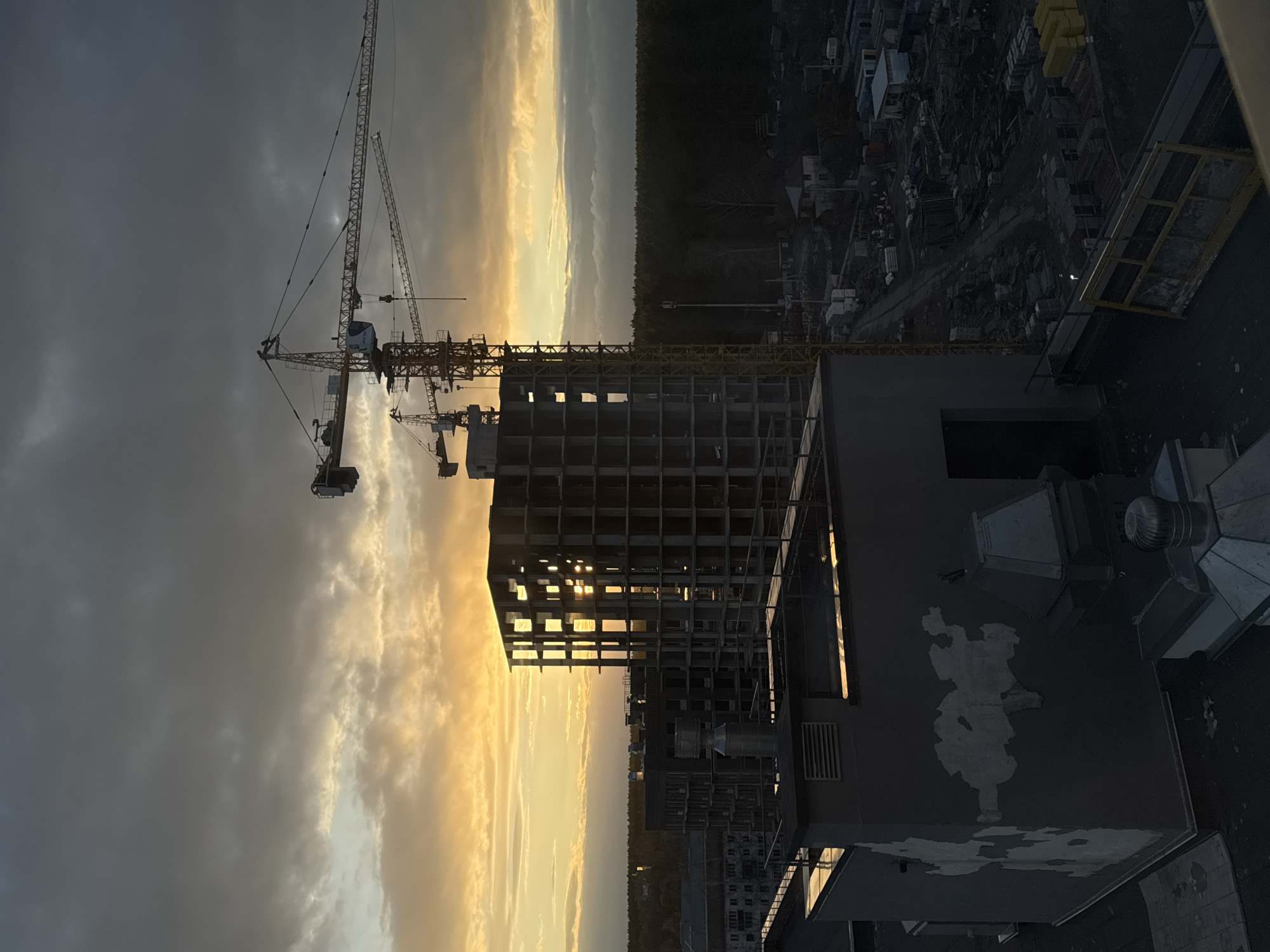 The image depicts a construction site at either sunrise or sunset in Izhevsk, Russia, viewed from an elevated perspective. In the foreground, the dark, flat rooftop of a building is visible on the right, featuring various industrial vents and equipment, and notable patches of peeling paint or concrete that form an irregular pattern.
Dominating the left and central midground are several towering construction cranes, silhouetted against a dramatic sky. One large tower crane is prominent on the left, with its jib extending across the frame. Behind it, another crane is partially visible. Adjacent to the cranes, a multi-story building under construction stands in a skeletal state, its concrete framework showing numerous empty window openings that catch the golden light of the horizon.
The sky is largely overcast with dark, heavy clouds, but a distinct band of bright golden and orange light stretches across the horizon, suggesting a low sun. This light bathes the distant clouds and the upper parts of the construction site. Below the bright horizon, a dense, dark line of trees or forest demarcates the mid-distance, separating the construction area from what appears to be a sprawling, dark landscape of further development or industrial ground in the far background.
The lighting creates a strong contrast, silhouetting many elements against the vibrant sky. The scene conveys an impression of urban expansion and industry during a transitional time of day. No people are visible, and no discernible text is clearly legible on any objects in the image.