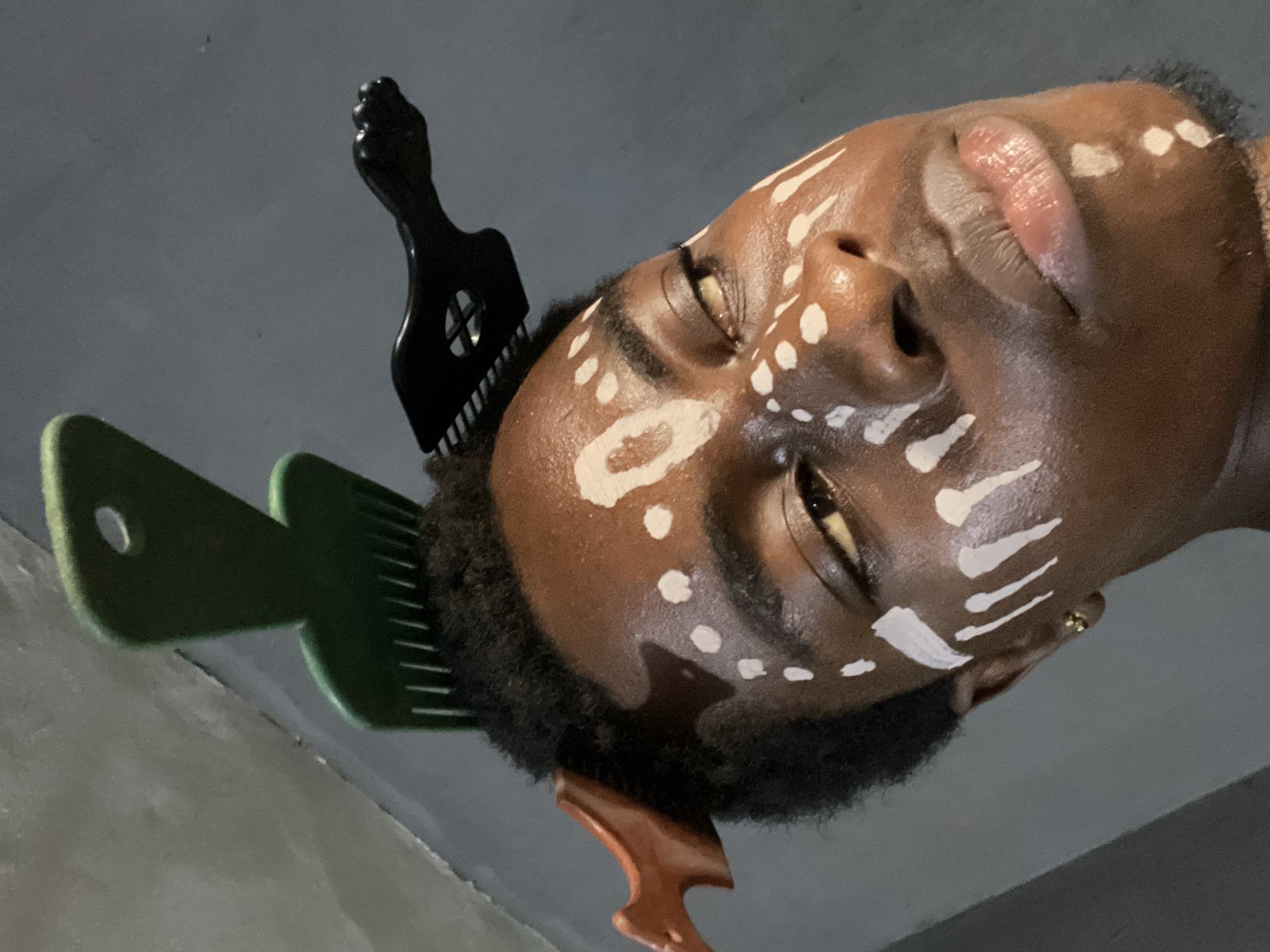The image shows a person with brown skin facing the camera. The person's face is adorned with white painted markings. Three hair picks are placed in the person's hair: one black, one green, and one brown. The background is a neutral gray color. The image does not provide information about the city of Nkpor, Nigeria.