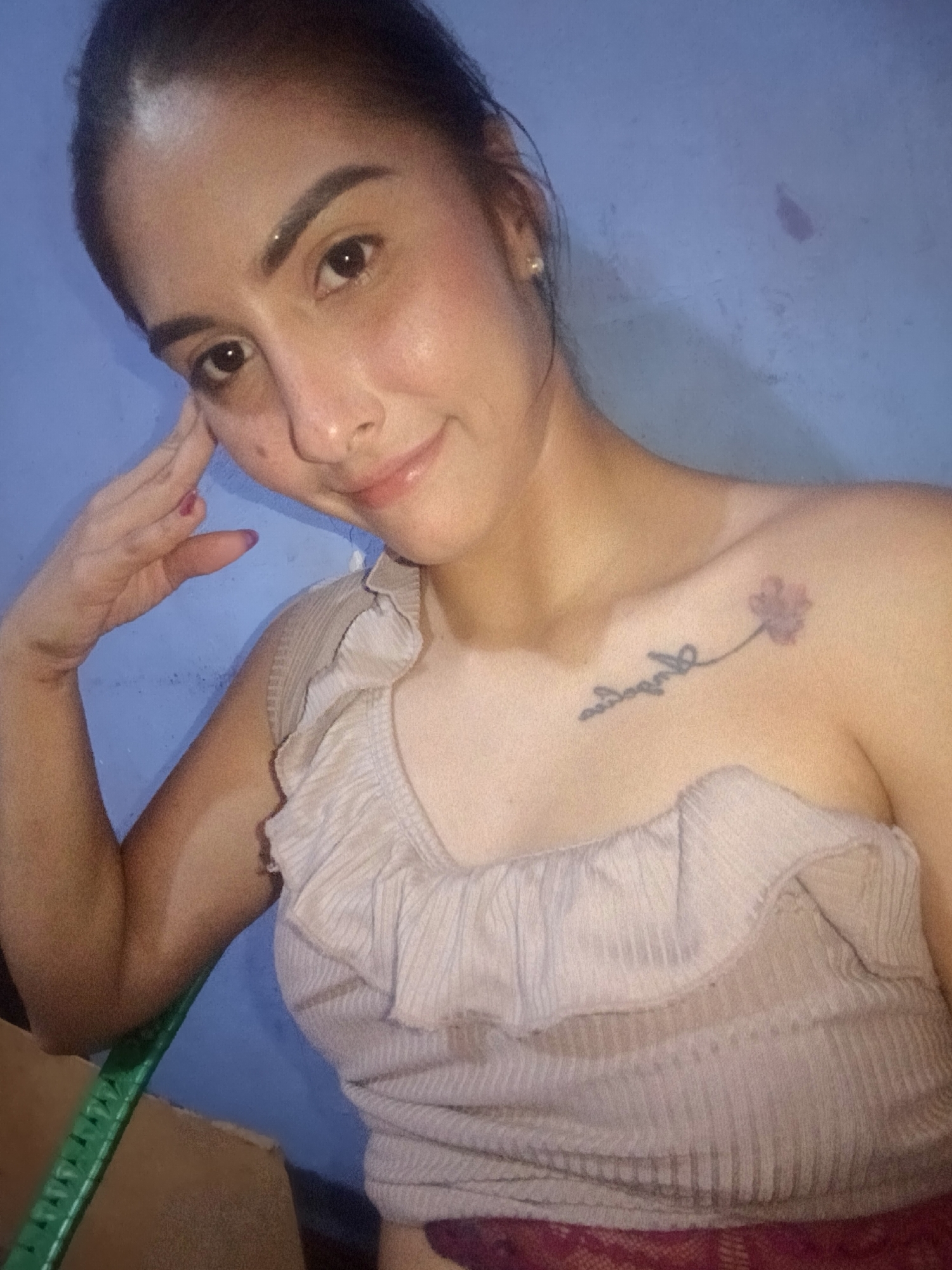 A fair-skinned woman is photographed indoors. She is resting her face on her left hand, which has purple nail polish. She is wearing a light brown, one-shoulder top with ruffled detailing. A tattoo of a flower and some script is visible on her right collarbone. The background is a light blue wall. The location in Juan Griego, Venezuela, cannot be determined from the image.