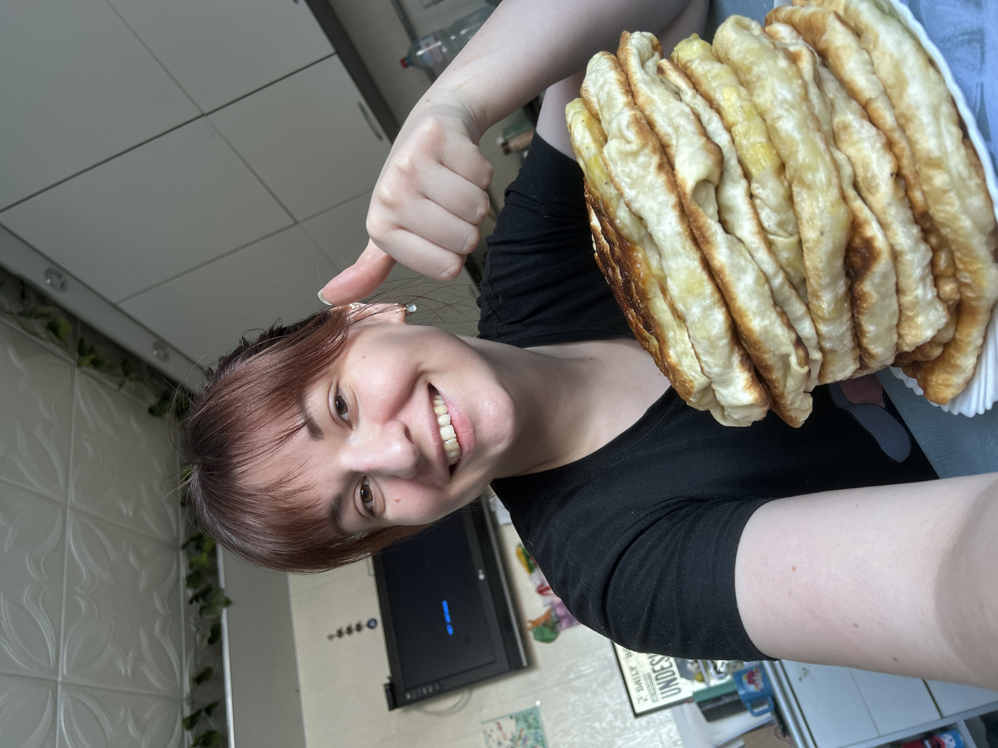 A woman is depicted in a close-up, selfie-style photograph, presenting a large stack of flatbreads or pancakes. She is smiling broadly and giving a thumbs-up gesture with her right hand. The woman has shoulder-length reddish-brown hair with bangs, light brown eyes, and is wearing a black t-shirt and small, light-colored earrings. Her left arm is partially visible on the lower left.
On a light blue surface in the foreground, a tall stack of approximately ten to twelve golden-brown, possibly fried or pan-cooked, flatbreads is arranged on a white plate.
The background suggests an indoor domestic setting, likely a kitchen or dining area, in the city of Фоки, Russia. To the left, a wall-mounted black LG television screen is visible, displaying "Нет сигнала" (No signal). Below the television, a light-colored dresser or cabinet holds various items, including a framed picture that appears to be a "Daily Prophet" newspaper prop and a blue mug. Above the television, a decorative item resembling a string of blue beads is hanging. The wall behind her is light-colored and textured. To the right, white overhead kitchen cabinets are visible, and behind her right shoulder, a plastic bottle stands on a counter. The ceiling above features decorative white panels, with some green foliage or decoration visible along the wall-ceiling junction.