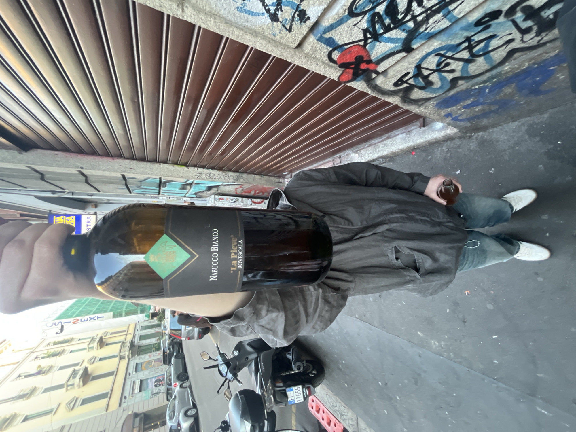 An outdoor, eye-level shot captures a person holding a bottle of white wine, with an urban street scene in the background. The image is oriented sideways, with the person on the right side of the frame.
The central subject is a person, largely visible from the waist down, wearing a dark jacket, blue jeans, and white sneakers. The person's right hand is holding a clear glass bottle of white wine, labeled "NABUCCO BIANCO" with "LA PIEVE" and "BOTTEGA ROVESCALA" below it, featuring a green diamond logo. The person's left hand is also holding another small, dark glass bottle, possibly a beer or cider bottle.
The background shows a street with buildings and parked vehicles. On the left, several multi-story buildings with balconies and window air conditioning units line the street. A bright blue sign with partially visible white text, possibly reading "YA" or "STRA" and "NEXT", is seen high on a building. A black scooter or moped with an Italian license plate "FL 11655" is parked on the street next to a red and white barrier. Several cars are also visible in the distance.
Above the person, there's a concrete overhang or wall covered in colorful graffiti, predominantly black, blue, and red tags. Below this wall is a brown, slatted roll-down security shutter, typical of storefronts. The ground is a dark asphalt or concrete surface.
The scene takes place during the daytime, indicated by the natural light. The weather appears clear. The overall setting is an urban environment, likely a European city given the scooter license plate and architectural style. No specific emotions are discernible from the partial view of the person.