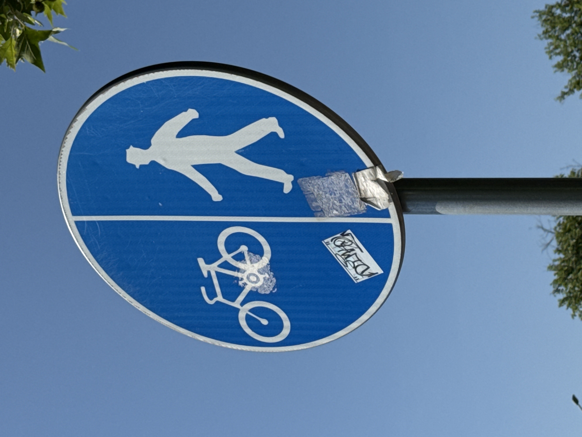 The image shows a blue, circular traffic sign divided vertically in half. The left side depicts a white silhouette of a bicycle, and the right side shows a white silhouette of a person walking. The sign is mounted on a grey metal pole. There are two stickers on the sign: one partially obscuring the bicycle and another on the right side, near the bottom. The sign is set against a clear blue sky, with some green foliage visible at the top and bottom edges of the frame. This sign indicates a shared path for cyclists and pedestrians.