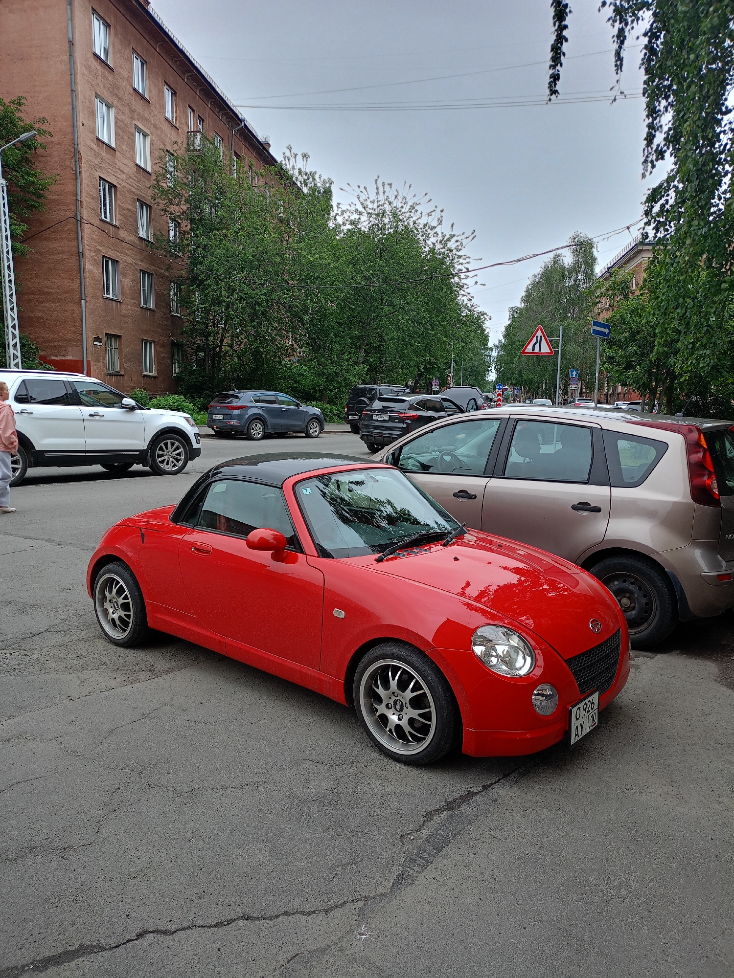 This media file depicts a street scene with several cars parked along the roadside and a person walking in the foreground. A bright red convertible is the most prominent object, parked in the center of the frame. To its left, a white SUV is visible. Behind the red car, a beige hatchback is parked. Further in the background, other vehicles and a multi-story building can be seen.
The scene appears to be set in an urban or suburban environment, with trees lining the street and a building that suggests residential housing. Traffic signs are visible, indicating a road with traffic regulations.
The time of day is unclear, but the overcast sky suggests it might be daytime with cloudy weather. There are no overt signs of activity beyond the parked cars and the pedestrian. The overall atmosphere is calm and ordinary, with no strong emotional cues conveyed.
Visible text includes license plates on the cars, such as "Q 926 AY 10" on the red convertible. The other license plates are either obscured or unreadable.