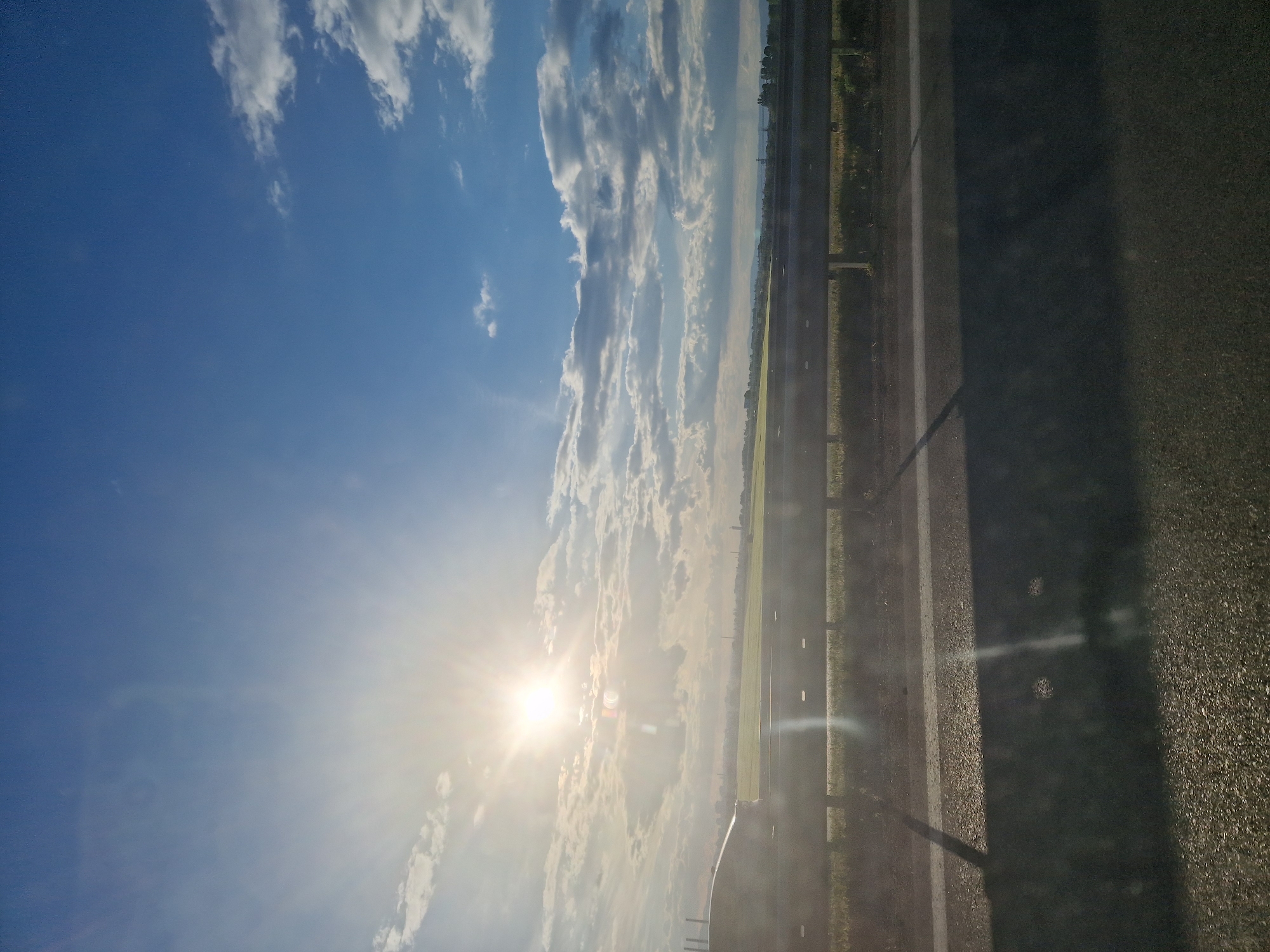 A daytime, low-angle shot depicts a road beside a field, with a bright blue sky above. Wispy, scattered clouds are present in the sky, and the sun is shining brightly through them. A road with white lines on the side stretches along the right side of the frame. The road is next to a green field. The location is in the city of Unknown, Unknown.