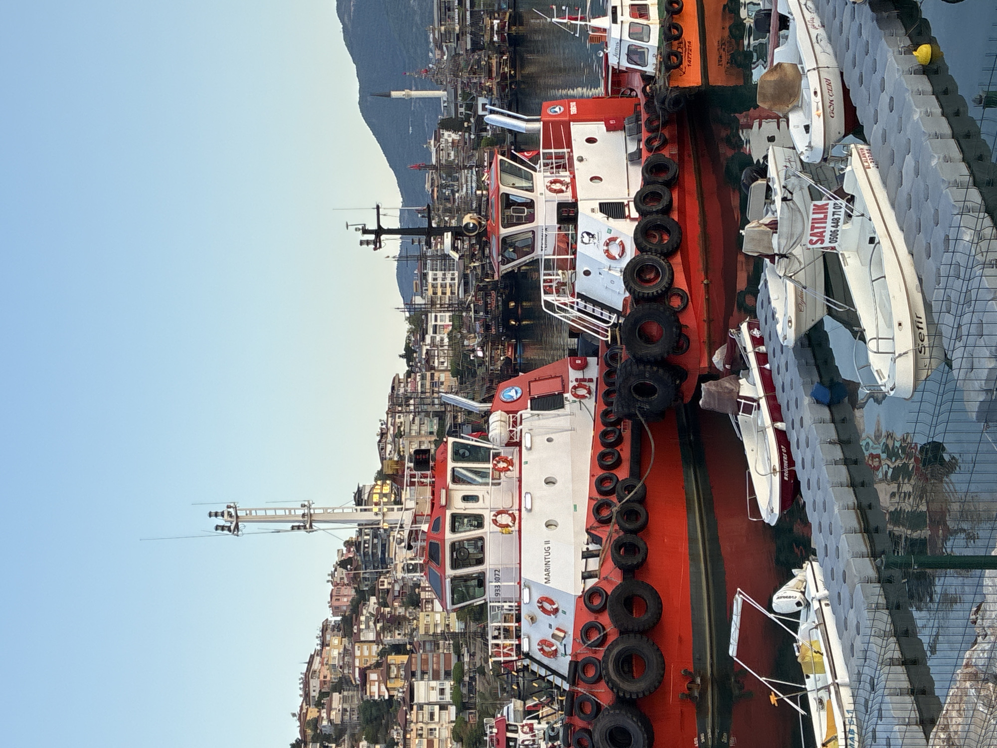 This image depicts a harbor scene in Alanya, Turkey, with several tugboats docked. In the foreground, two prominent red and white tugboats, identified as "MARINTUG II" and another with a number "933 3072" on its superstructure, are prominently featured. Both vessels are equipped with numerous black tires along their sides, serving as fenders. Smaller white boats are also visible, docked alongside floating pontoons in the lower portion of the image. In the background, a hillside covered with densely packed buildings, typical of Mediterranean coastal towns, rises steeply. Further in the distance, a minaret is visible, indicating a mosque, and more boats are moored in the harbor. The sky is clear and blue, suggesting a daytime scene. There are no people clearly visible in the image.