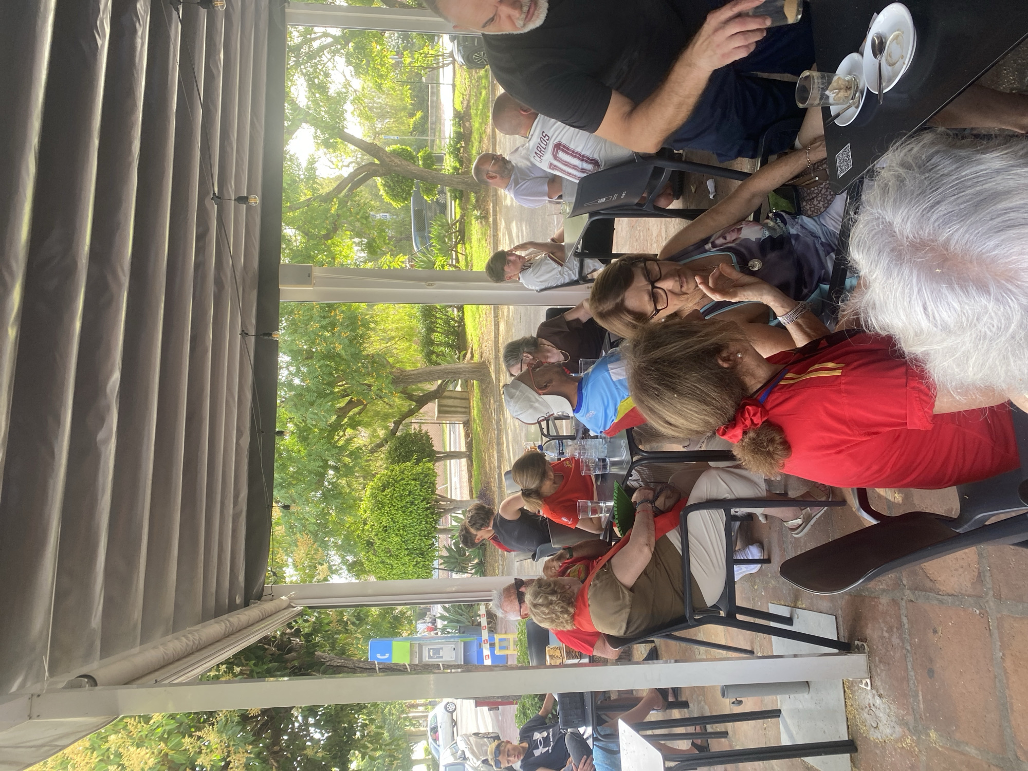 The image shows an outdoor seating area of a cafe or restaurant in San Pedro De Alcántara, Spain, likely during daytime, indicated by the bright sunlight filtering through the trees and awning. Several groups of people are seated at tables, engaged in conversation or consuming drinks and snacks. The environment includes a covered patio with a grey fabric awning supported by white pillars, and paved flooring. Lush green trees and foliage are visible in the background, suggesting a pleasant outdoor setting.
In the foreground, two women are seated at a black table. One woman, wearing a red top, is on the left, and the other, in a patterned top and glasses, is on the right. A person's head with grey hair is visible in front of them, suggesting they are a third person at the table. A QR code is visible on the table.
To the left, another group is seated. A man with white hair and sunglasses, wearing a red scarf, is in the center. Next to him, a woman in a brown top is seated. Further left, a young person with blonde hair is seated at another table.
In the middle ground, more people are seated at tables. A man wearing a white t-shirt with "CARLOS 10" printed on the back is visible. A person in a blue and red outfit is also seated.
In the background, a blue and yellow telephone booth is visible on the left, and cars are parked or driving on a street. The overall scene depicts a relaxed social gathering at an outdoor cafe.